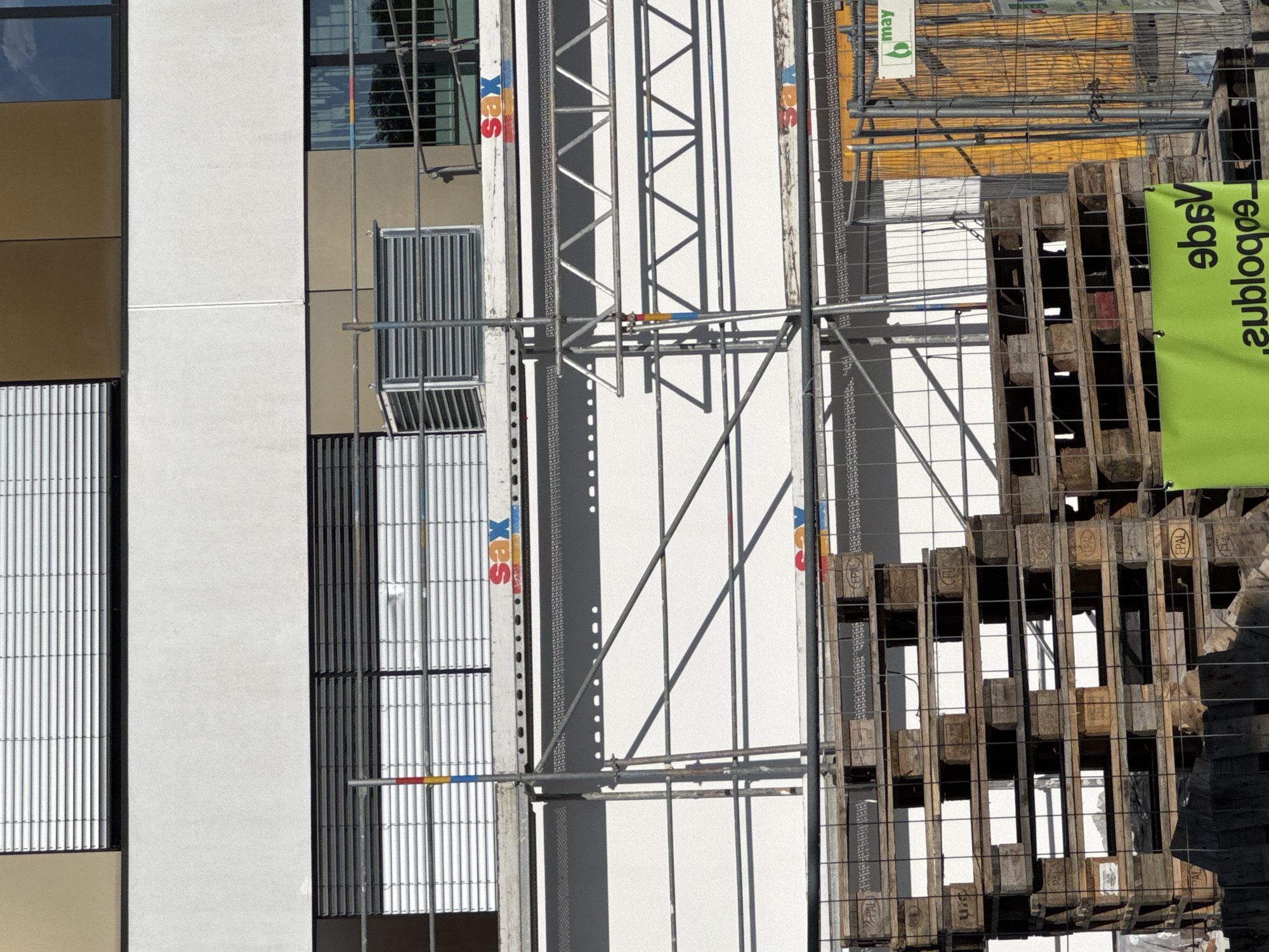 This is a photograph taken in Höhenkirchen-Siegertsbrunn, Germany, depicting a construction site. The scene is dominated by the side of a modern building with a beige and white facade, featuring large windows and vertical metal grating. Adjacent to the building is scaffolding, constructed from metal poles and bars, supporting a white panel that is likely part of the building's exterior.
On the right side of the image, a stack of wooden pallets is visible, secured by a metal mesh fence. Some of the pallets have the "EPAL" logo. A bright green banner with black text is draped over the pallets, displaying the words "eubloqna" and "9bsv" in a mirrored, vertical orientation. Above the pallets and scaffolding, there is a yellow construction barrier, and a white sign with a green leaf logo and the word "may" is attached to it.
The image appears to be taken during daylight, with strong shadows cast by the scaffolding onto the white panel, suggesting it is a sunny day. The overall impression is one of ongoing construction and development. There are no people visible in the frame. The text present includes "EPAL," "eubloqna," "9bsv," and "may."
