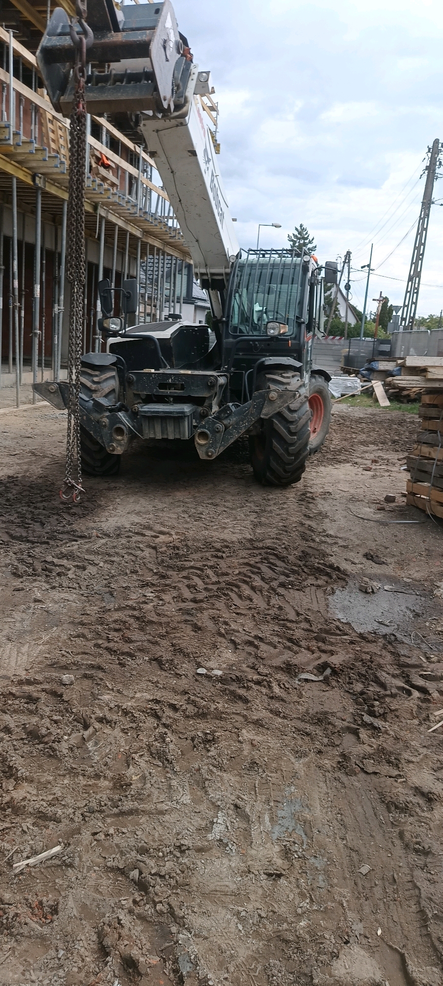 A construction site in Opole, Poland, is depicted in the image. A large, black telescopic handler, likely a Bobcat, is positioned in the foreground. The machine has a heavy-duty boom extended upwards and a chain hanging from its front attachment. The ground is muddy and marked with tire tracks, indicating recent activity. In the background, unfinished building structures with scaffolding are visible, along with piles of wood and construction materials. Power lines and a few buildings in the distance suggest an urban or semi-urban environment. The sky is overcast, with a few scattered clouds. No people are clearly visible in the scene.