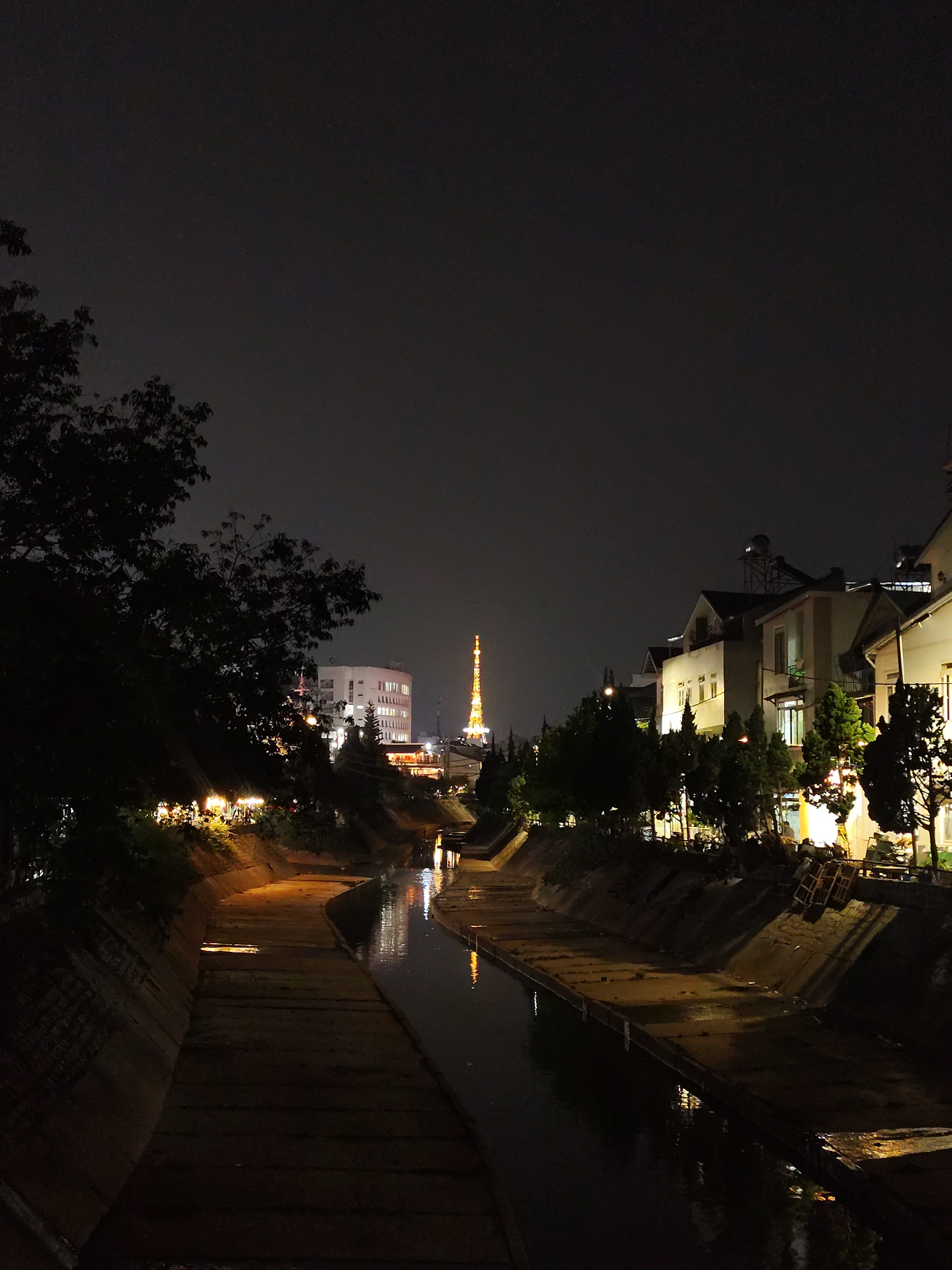 This nighttime photograph showcases a serene urban scene in Da Lat, Vietnam, centered around a concrete-lined canal. The dark sky dominates the upper portion of the image, contrasting sharply with the various artificial lights illuminating the cityscape below.
In the foreground, the canal's concrete banks, which are partially damp, recede into the distance. A narrow stream of water flows through the channel, reflecting the bright lights from the surroundings and creating shimmering streaks on its surface. To the left, the dense, dark foliage of a large tree occupies a significant part of the frame, obscuring some of the canal bank. Beyond the tree, scattered lights suggest activity along the waterway.
On the right side of the canal, a row of buildings, possibly residences, are brightly lit from within, their windows casting a warm glow. These structures feature pitched roofs and are interspersed with lush evergreen trees. In the mid-distance, a prominent, brightly illuminated tower, distinctly resembling the Eiffel Tower, rises against the dark backdrop, serving as a clear landmark. The overall impression is one of a quiet city night, with the canal acting as a central vein through the illuminated urban landscape. No visible text or specific human activity is discernible, contributing to the tranquil atmosphere.