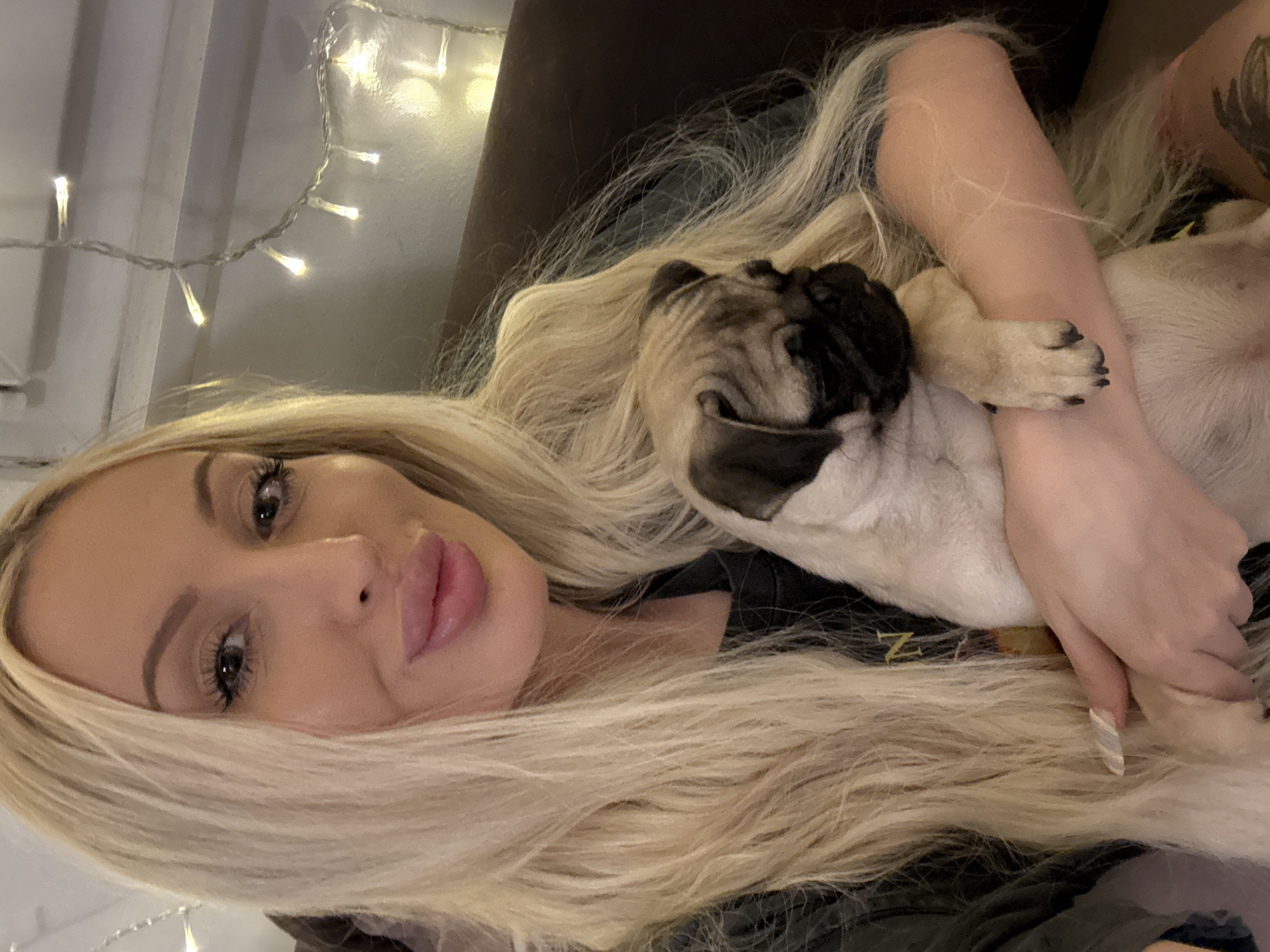 A woman with long blonde hair holds a pug puppy in her arms, looking at the camera with her lips pursed. The background is dimly lit with string lights. The woman is wearing a dark shirt. The pug puppy is light brown with black markings.