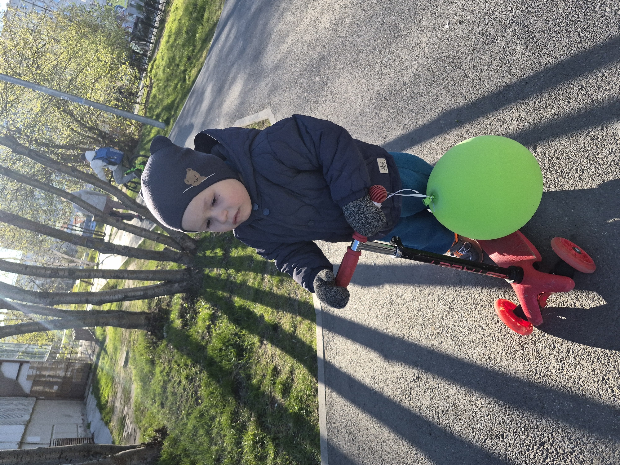A toddler, wearing a dark blue quilted jacket, a gray beanie with a bear emblem, and gray mittens, stands on a red scooter with a green balloon attached to it. The location appears to be a park or public space in Tobolsk, Russia, based on the architectural styles visible in the background. The scene is bathed in sunlight, casting shadows from the trees and the scooter onto the asphalt path. The toddler is facing downward, looking at the scooter. The setting suggests a warm, sunny day outdoors.