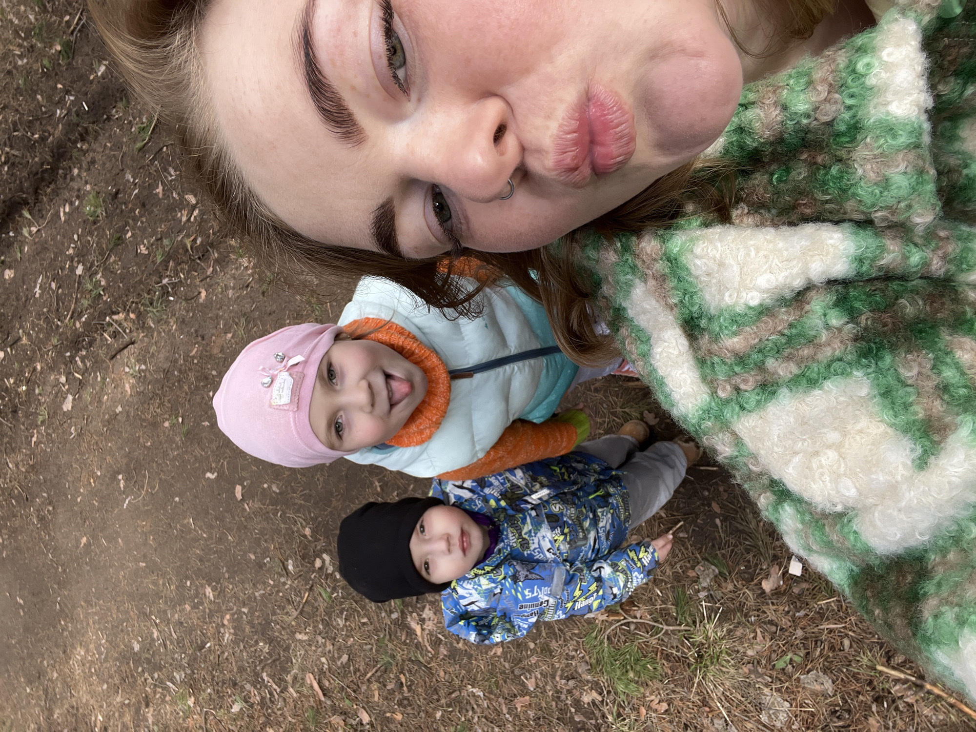 A selfie taken at ground level depicts a fair-skinned adult woman with a nose ring pouting directly into the camera. Below her are two young children, one wearing a pink hat, light blue jacket, and orange undershirt, sticking their tongue out, and another child wearing a black beanie and blue patterned jacket. The scene is set on a dirt and grass ground, possibly in a park. A green and white patterned soft garment covers part of the woman's right side. The location is likely in Chaikovsky, Russia.