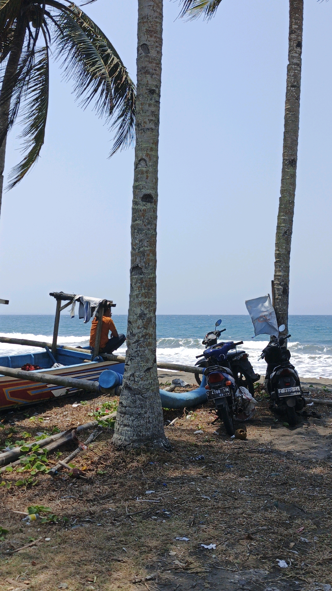 The image depicts a serene beach scene in Desa Batukaras, Indonesia, bathed in bright sunlight under a clear blue sky. In the foreground, two tall coconut palm trees stand prominently, their trunks textured and weathered. Between them, a wide expanse of blue ocean stretches to the horizon, with gentle waves lapping against the sandy shore.
On the beach, a traditional blue and orange fishing boat rests partially on the sand, its wooden structure weathered and displaying colorful painted details. A person, dressed in an orange shirt and dark pants, sits on the edge of the boat, facing the ocean and appearing contemplative. Clothes are draped over a wooden frame attached to the boat.
To the right of the boat and near the palm trees, three motorcycles are parked on the sandy ground. Two are dark-colored, and one is blue. They are positioned close together, suggesting they belong to the individuals present at the beach. A light-colored flag on a pole flutters gently in the breeze near one of the motorcycles.
The ground is a mix of sand, dry leaves, twigs, and some sparse green vegetation, indicating a natural, somewhat untamed beach environment. Debris, including small pieces of plastic and other litter, is scattered across the ground, hinting at human activity. The overall atmosphere is one of a peaceful, coastal resting spot.
