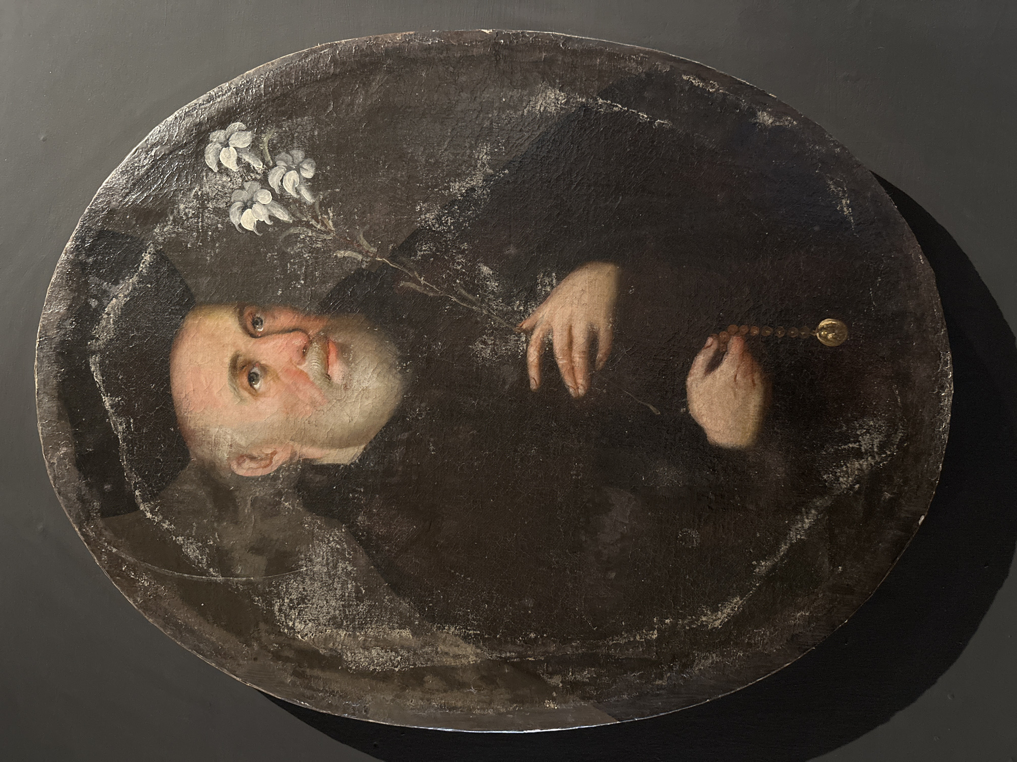 The image shows an oval-framed portrait of a man with fair skin, a bald head, and a slight blush to his cheeks. He is wearing dark clothing and is holding a rosary in one hand and a white flower sprig in the other. The painting appears old and has noticeable wear and tear. The background behind the painting is a solid dark color. There is no information related to the location in San Miguel Zinacantepec, Mexico.