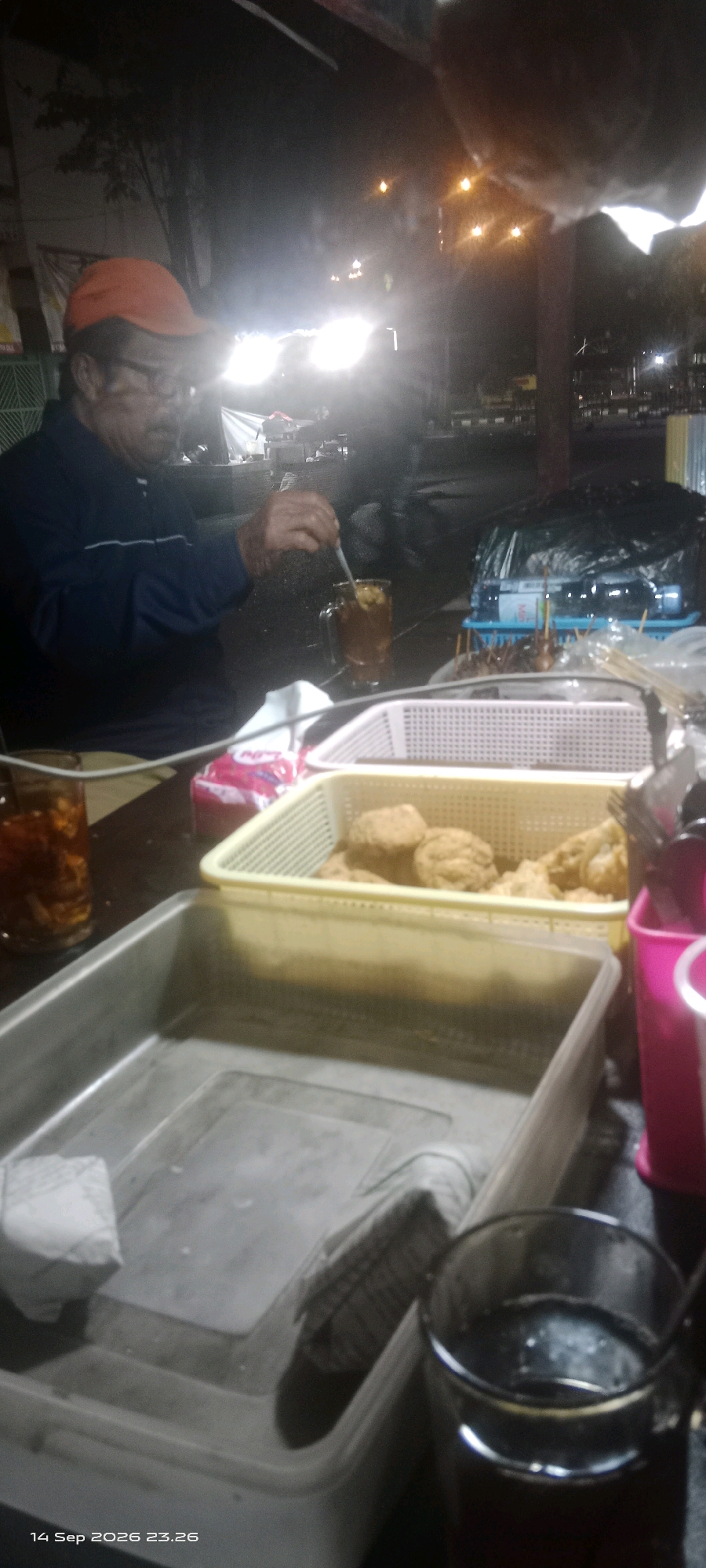 An outdoor night scene in Klaten, Indonesia, shows an elderly man wearing a blue jacket and an orange cap. He is seated at a table, stirring a glass of iced tea with a spoon. In front of him are two plastic containers filled with fried food, possibly tempeh or tofu, and a larger metal container. A second glass of iced tea is visible to his left. The background is dimly lit with artificial lights and shows some vegetation and structures, suggesting a street food stall or a roadside eatery. The time stamp on the image indicates September 14, 2026, at 11:26 PM.