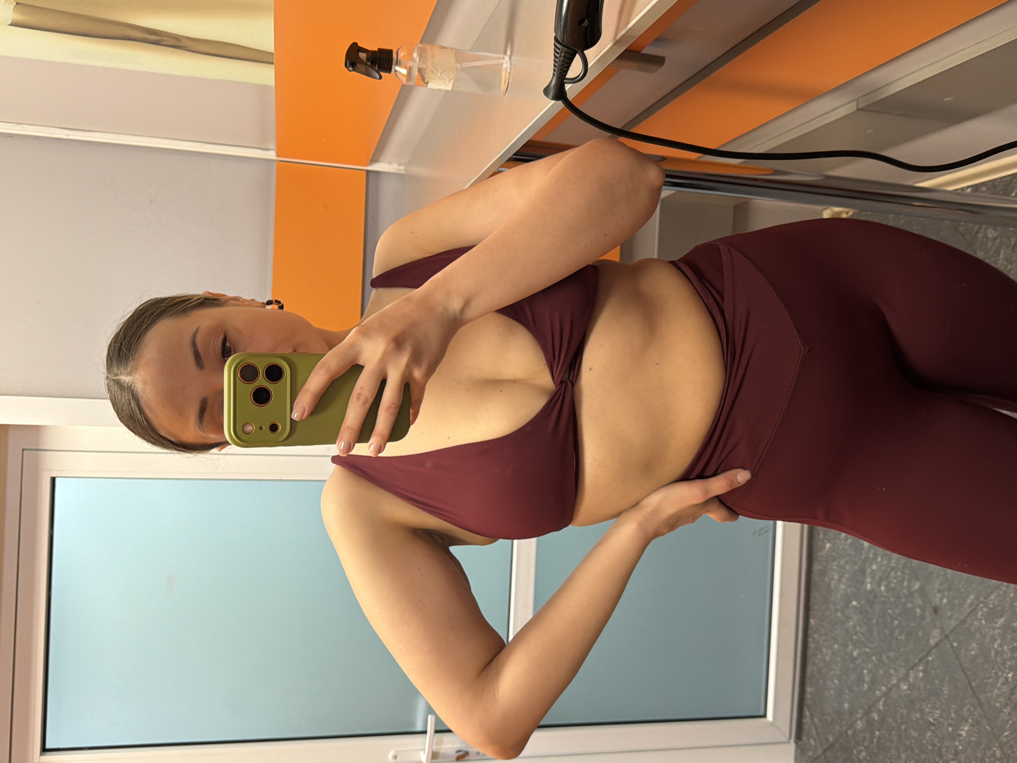 A woman is taking a selfie in front of a mirror. She is wearing a maroon-colored athletic bra and leggings. She holds a green smartphone in her left hand, partially obscuring her face. The background includes a window with frosted glass, a table with orange and light grey panels, a spray bottle, and a hair dryer with a black power cord. The floor is grey with a speckled pattern. The location may be a gym or spa, but there is no definite indication in the image. The city of Krasnoyarsk, Russia cannot be verified from the image.
