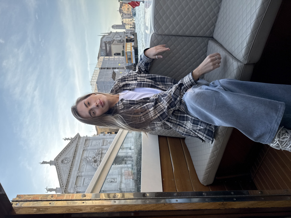 A fair-skinned adult female, with long blonde hair parted to her right, is seated and looking directly at the camera with a subtle smile. She wears a white crew-neck t-shirt, an unbuttoned grey, black, and white plaid long-sleeve shirt, light blue wide-leg jeans with a small tear on the left thigh, and white athletic shoes. Rings are visible on her fingers. She occupies a light grey, quilted bench on the left side of a boat, which features polished wooden paneling.
The boat is positioned on a wide canal in Venice, Italy. In the immediate background, on the left bank of the canal, the ornate Baroque facade of the Church of San Stae (Chiesa di Sant'Eustachio) is prominently visible, featuring statues on its roofline and a decorative central entrance. To the right of the church, several buildings with scaffolding indicate ongoing construction or renovation. Further down the canal, additional Venetian architecture and a distant campanile are observable under a partly cloudy sky. The water in the canal shows slight ripples.