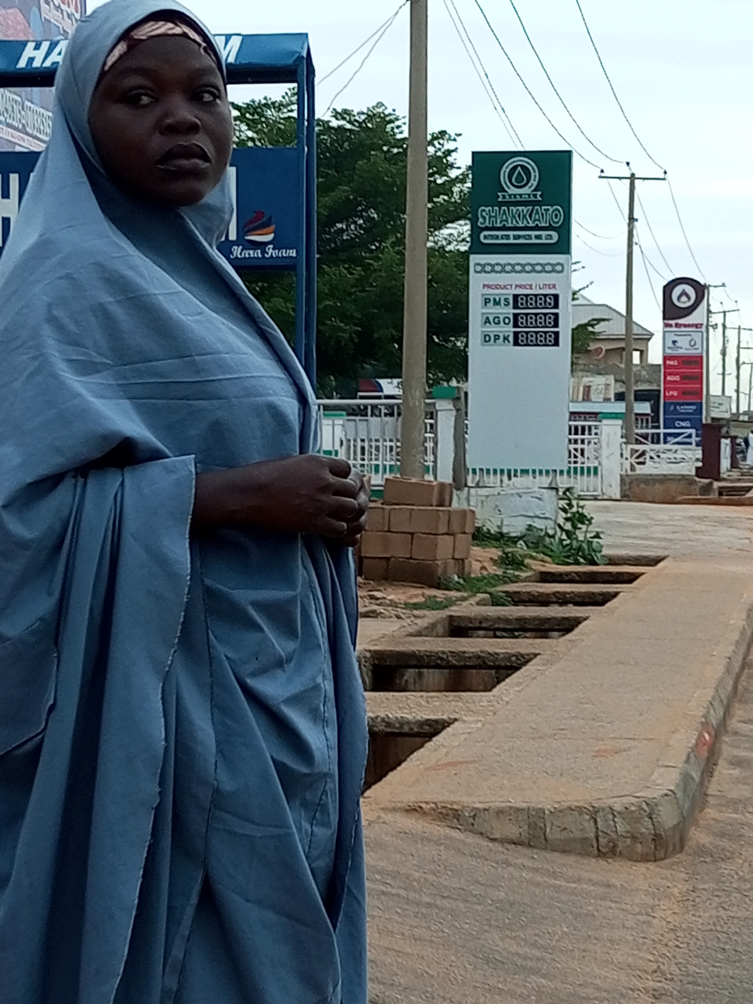 In Garko, Nigeria, a person wearing a blue hijab and a matching flowing garment stands in front of a fuel station. The person's face is visible, with dark skin and dark eyes, and they are looking to the left. Behind them, a tall, rectangular sign displays "SHAKKATO" at the top in green lettering, followed by "PRODUCT PRICE / LITER" and then a list of fuel types with prices: "PMS 8888", "AGO 8888", and "DPK 8888". To the right of this sign, another, shorter sign displays fuel prices in red. In the background, there are trees, buildings, and power lines. The ground in the foreground is a mix of dirt and concrete, with a drainage ditch visible. The scene appears to be an outdoor area at a fuel station, likely during the daytime due to the lighting.