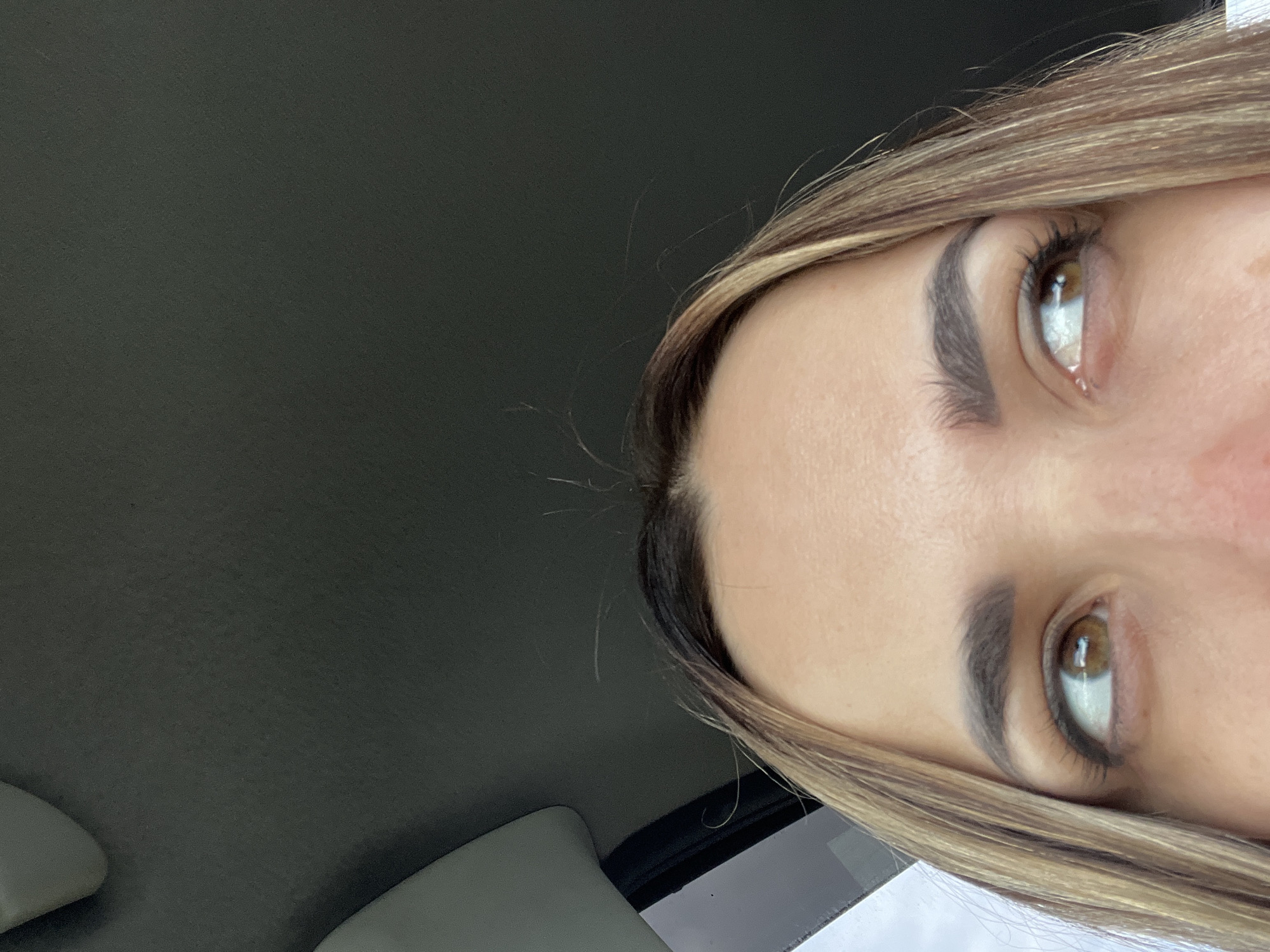 A close-up image displays the upper portion of a female subject's face, including her forehead, eyebrows, and eyes. She has light brown hair with blonde highlights, parted centrally, and her brown/hazel eyes are directed towards the upper right of the frame. Her eyebrows are dark and defined. The background features the dark gray, textured headliner of a vehicle, with a lighter gray interior trim piece and a window frame partially visible on the upper left, revealing a glimpse of an overcast exterior. The subject is seated within a vehicle. The location is an unknown city, Unknown.