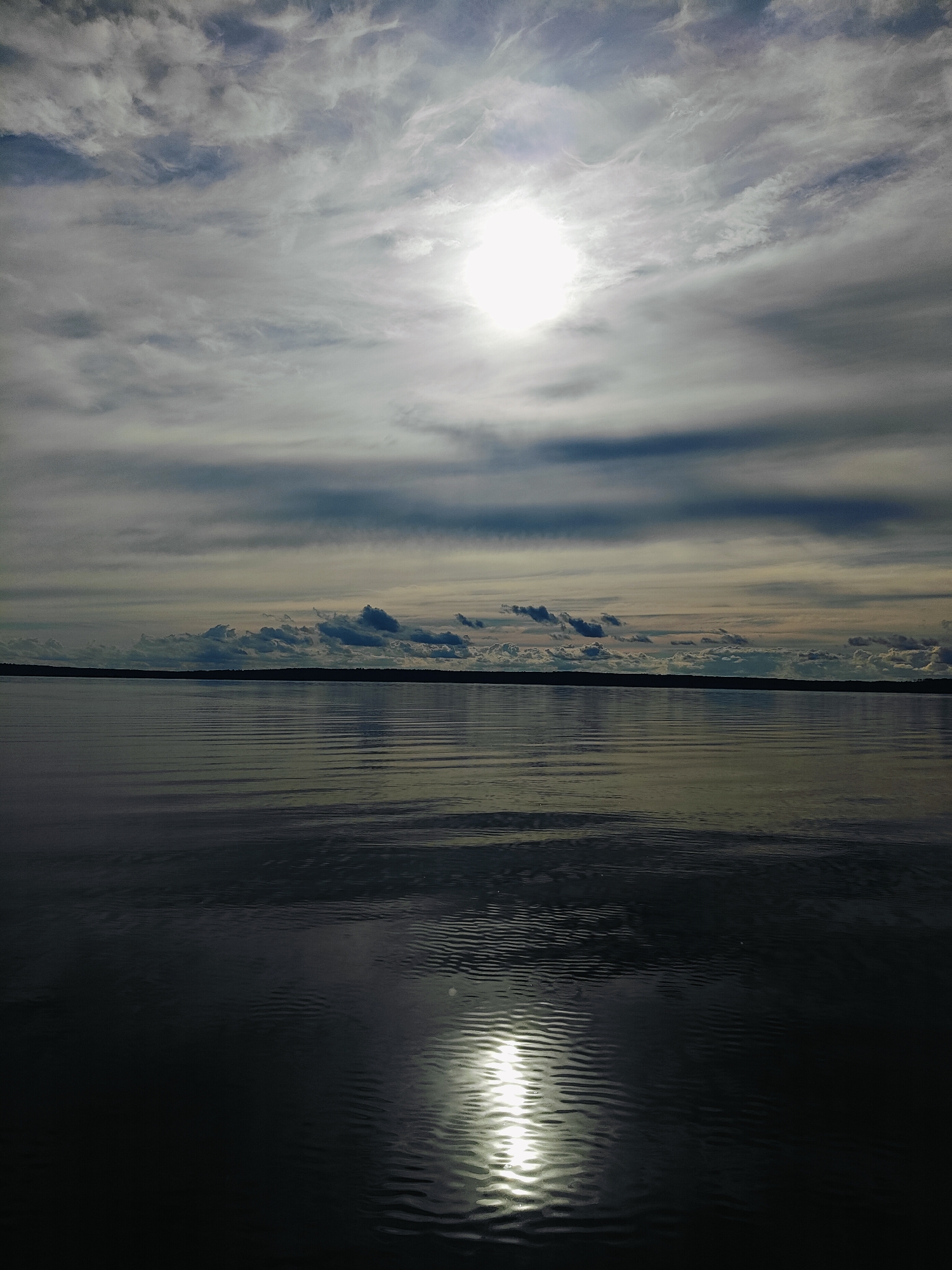 A vast, dark body of water stretches towards a distant, dark tree line under a cloudy sky. The sun shines brightly through the clouds, casting a shimmering reflection on the water's surface, creating ripples and waves. The clouds are wispy and layered, with some darker, more defined clouds scattered amongst them. The overall mood is serene and contemplative, with the natural elements dominating the scene. The location is Stepanovo, Russia. There are no people or animals visible in the image.