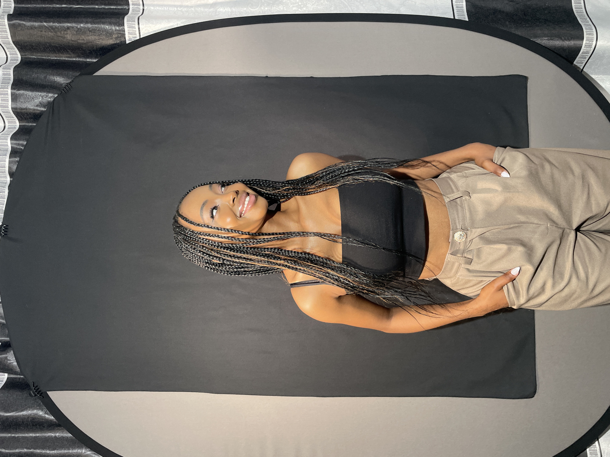 This media file captures a portrait of a young Black woman in a studio setting, photographed from an overhead perspective. She is lying horizontally in the frame, with her head positioned to the left.
The woman has long, intricately braided black hair that frames her face and spreads around her head. She is smiling brightly, revealing her teeth, and her eyes are open, looking slightly upwards, conveying a happy and confident demeanor. Her makeup highlights her eyes and lips. She is dressed in a black strapless crop top, exposing her midriff, paired with light brown or khaki high-waisted pants that feature a decorative button on the waistline. Her fingernails are painted with white polish, visible on her hands that rest near her waist.
The setting appears to be a photography studio. The woman is reclining on a large, circular mat. The central area where she lies is a rectangular section of dark gray or black fabric, surrounded by a lighter gray circular border, which is edged by a black rim. Beyond this mat, the background features a textured surface, possibly a corrugated studio backdrop. The left side of the background displays a black and white striped pattern, while the right side shows a more subtly patterned, lighter gray texture.
The image suggests a professional photoshoot, likely conducted indoors with controlled lighting, focusing on the model's joyful expression and stylish attire. No visible text is present in the image.