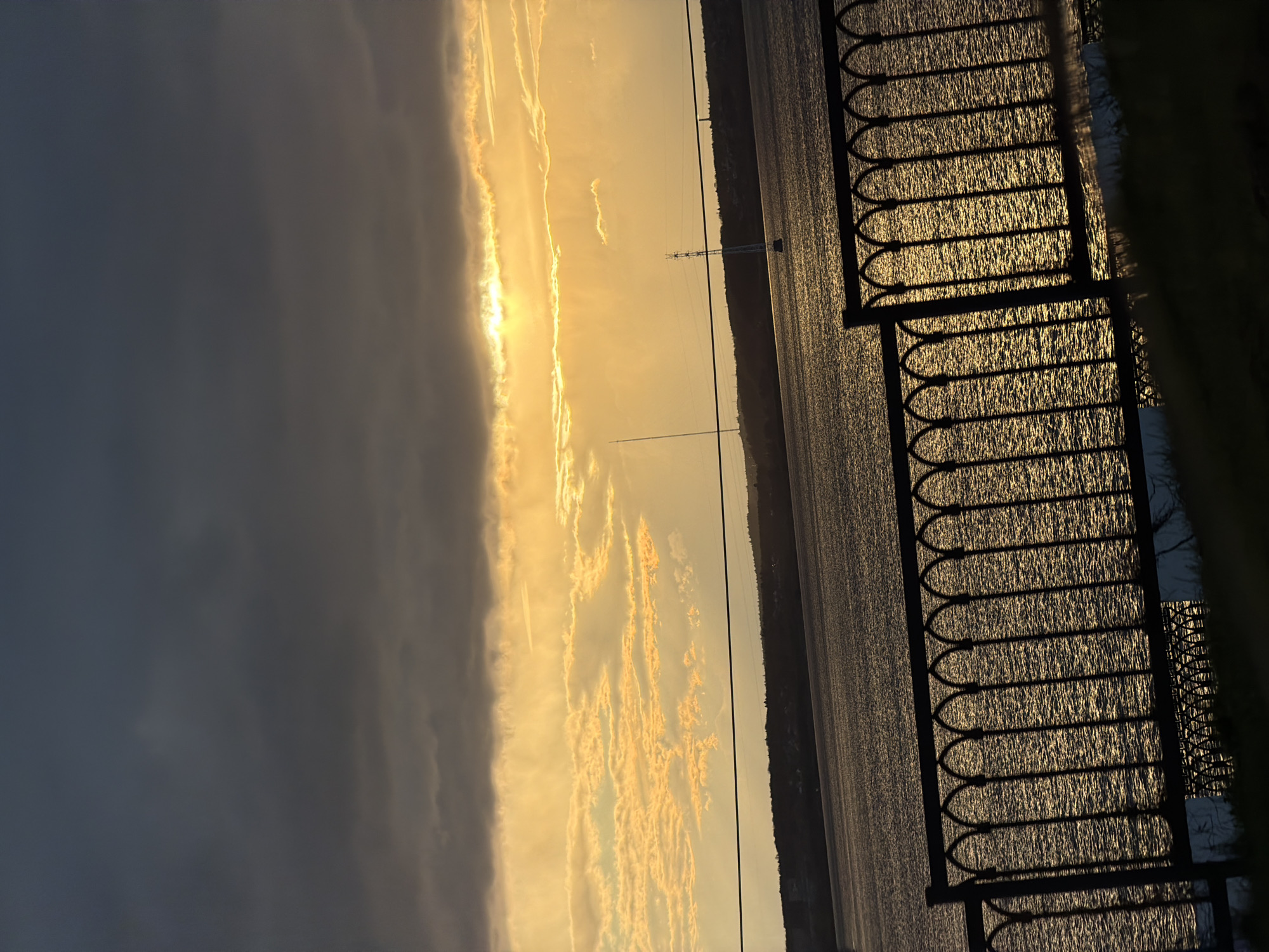 The image captures a vibrant sunset or sunrise over a large body of water, likely a river or lake, viewed from an embankment. The scene is dominated by warm, golden light emanating from the horizon.
In the foreground, a dark, ornate metal fence with a repeating arched pattern is silhouetted against the bright, shimmering water. The surface of the water reflects the golden light intensely, creating a textured, glittering effect. A hint of dark green foliage is visible on the far right edge, possibly indicating a grassy area or bushes behind the fence.
The midground features the expansive water body, stretching towards a distant, low-lying landmass on the horizon. A tall, slender mast or utility pole stands prominently in the water, appearing as a dark vertical line against the illuminated sky and water.
The sky is a significant element, divided into contrasting sections. On the left, a large mass of dark, heavy grey clouds creates a moody atmosphere, contrasting sharply with the right side of the sky, which is aglow with brilliant golden and orange hues. Here, streaky and broken clouds are dramatically lit by the low sun.
The time of day is clearly either sunset or sunrise, characterized by the strong, low-angle golden light and long shadows. The overall mood is serene and visually striking due to the stark contrasts and vivid colors. The location context provided indicates this scene is in Izhevsk, Russia. There is no visible text in the media file.