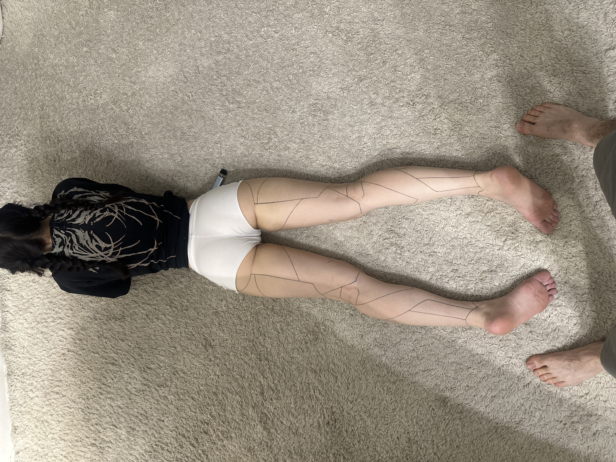 A person lies prone on a beige carpet with geometric lines drawn on their legs. The person has dark hair in braids and wears a black shirt with a white pattern and white shorts. A second person's bare feet are visible near the first person's feet. The location is unspecified. The scene lacks contextual indicators suggesting it is in Khimki, Russia.