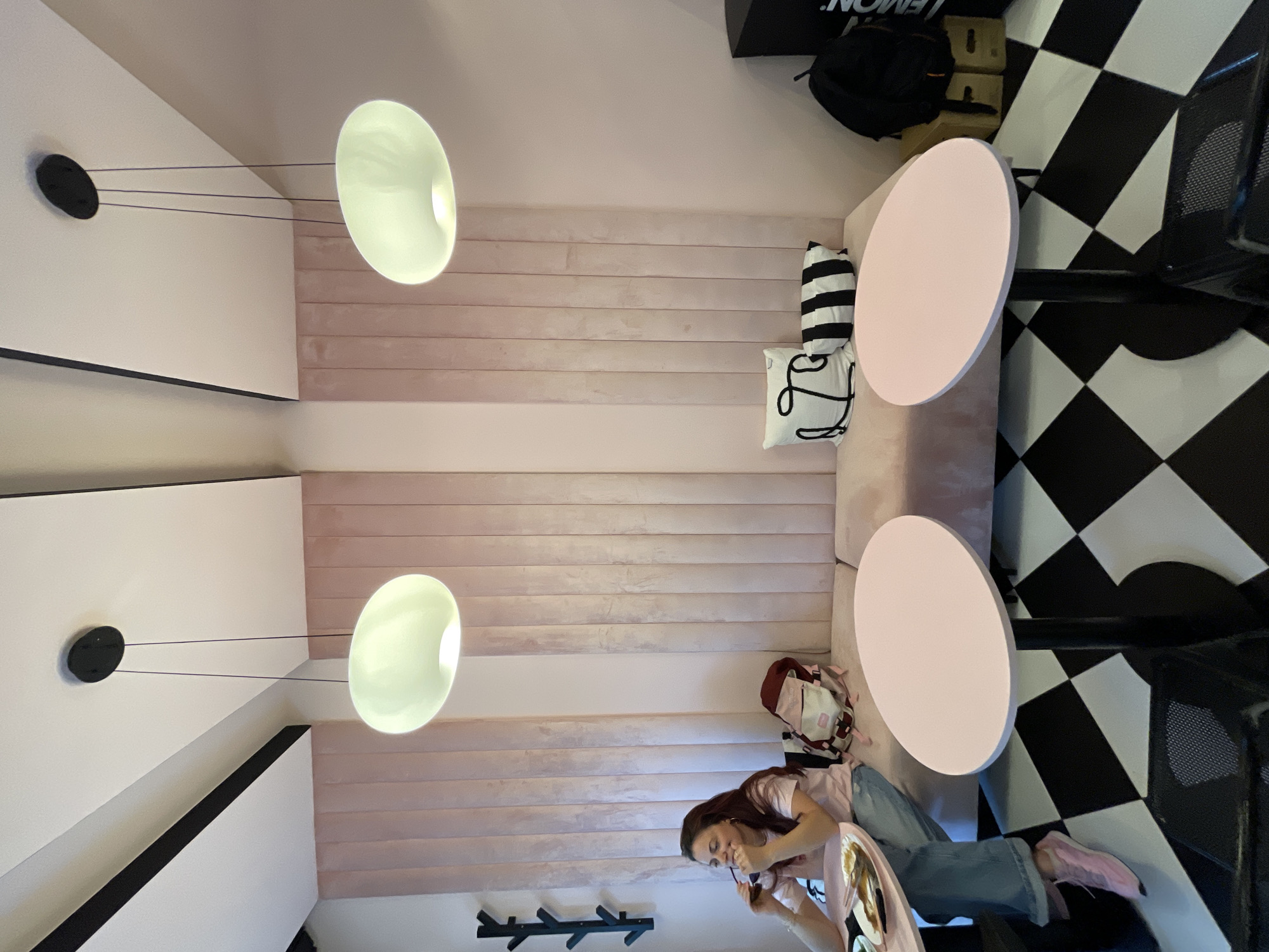 The image depicts an indoor scene within a modern cafe or restaurant located in Sofia, Bulgaria. The environment features a distinct interior design characterized by soft pink and white tones contrasted with black elements.
The left and central walls are primarily covered with vertical panels of pink velvet upholstery, extending from mid-height upwards. Above these panels, the walls are white. The rightmost visible wall section is a smooth, plain pink. The ceiling is white, traversed by two prominent parallel black structural beams. From the white ceiling, two oval-shaped pendant lights, emitting a pale green-white glow, hang on slender black wires from black circular mounts.
The floor is covered in a large black and white checkered pattern. Along the upholstered pink wall, a continuous, low pink upholstered bench or sofa serves as seating.
On the left side of the frame, an adult female is seated on the pink bench at a round, light pink table with a black pedestal base. She has long, reddish-brown hair and is wearing a light pink t-shirt and blue jeans (visible below the table). Her feet, adorned in pink sneakers, rest on the checkered floor. She is looking towards the right with a slight smile, holding a pair of dark sunglasses near her mouth with her left hand. On the table in front of her is a white plate containing remnants of food, including what appears to be fish bones and sauce. A fork and knife are resting on the plate. A dark beverage in a glass is partially visible next to the plate. To her immediate right, a maroon and white backpack rests on the pink bench.
Further to the right, a second identical round, light pink table with a black pedestal base stands unoccupied. On the pink bench between the two tables, two decorative pillows are placed: one white with a black abstract linear design, and one black with white horizontal stripes. In the far right background, a black audio speaker or box is visible, stacked on top of several plain beige boxes. A black backpack rests on these boxes. On the far left wall, a black, multi-pronged coat rack resembling a tree branch is mounted.