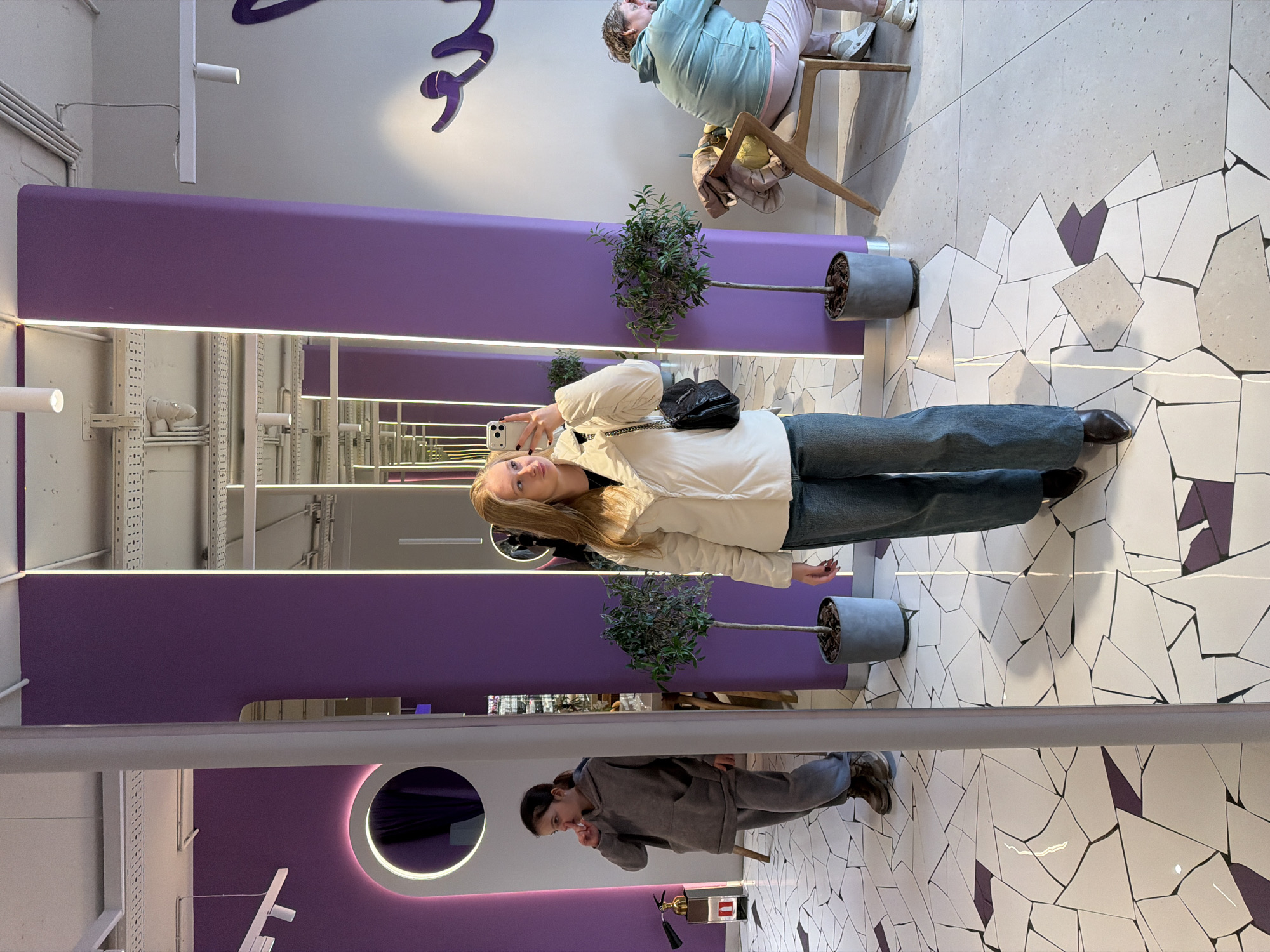 A woman stands in a modern, indoor space. She is fair-skinned with long blonde hair. She wears a white jacket, dark top, blue jeans, and dark shoes. She is holding a cell phone in her left hand, as if taking a selfie, and has a small black purse over her left shoulder. Her right arm is extended slightly to the side. She is positioned near a potted tree.
The environment is characterized by a tiled floor with irregular, angular patterns in white, gray, and purple. There are purple rectangular structures with horizontal lighting. Above these structures is exposed structural framework. Another person is visible in the reflection of a mirror-like surface below, dressed in a gray outfit. A third person is seated on a stool in the upper right corner of the frame, also reflected.
The location may be in Krasnodar, Russia.