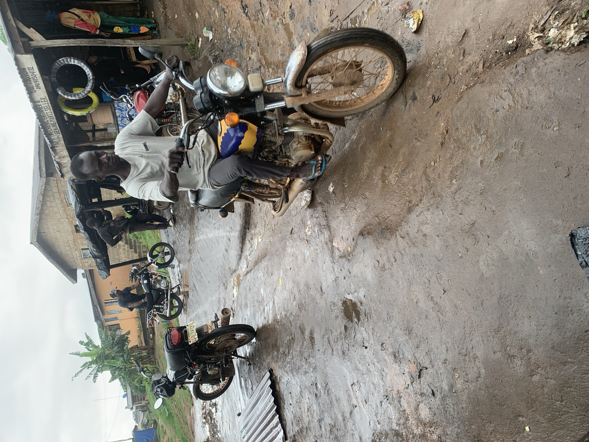 A medium shot captures a man riding a motorcycle down a muddy, wet street in Shagamu, Nigeria. The man, wearing a light-colored t-shirt and dark pants, is in focus, with the motorcycle's front wheel slightly turned. The street is characterized by puddles and dirt, suggesting recent rainfall. In the background, a few other motorcycles are parked, and a small shop with a corrugated metal roof is visible. Two other individuals are present in the background, one standing near the shop and another leaning against a motorcycle. The overall scene depicts a typical street view in a developing urban environment.