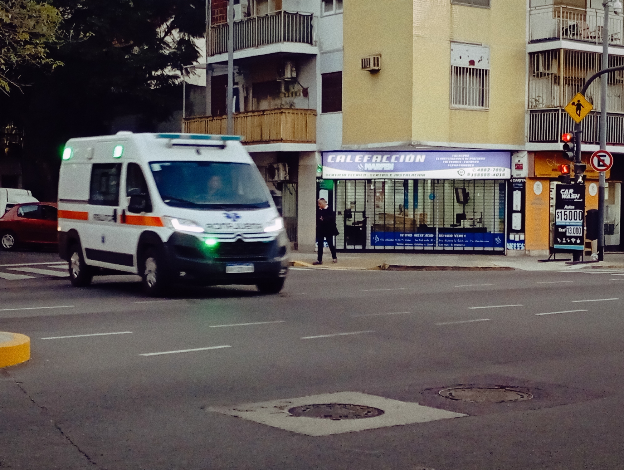 The image captures a street scene in Buenos Aires, Argentina. An ambulance with illuminated green lights is moving on the road. A pedestrian is seen standing on the sidewalk near a building. The building has a storefront with "CALEFACCIÓN SHARPEN" signage. A traffic light is visible with a red signal. Next to the traffic light is a sign advertising a "CAR WASH" with prices. The street has lane markings and manhole covers. Buildings with balconies are present in the background.