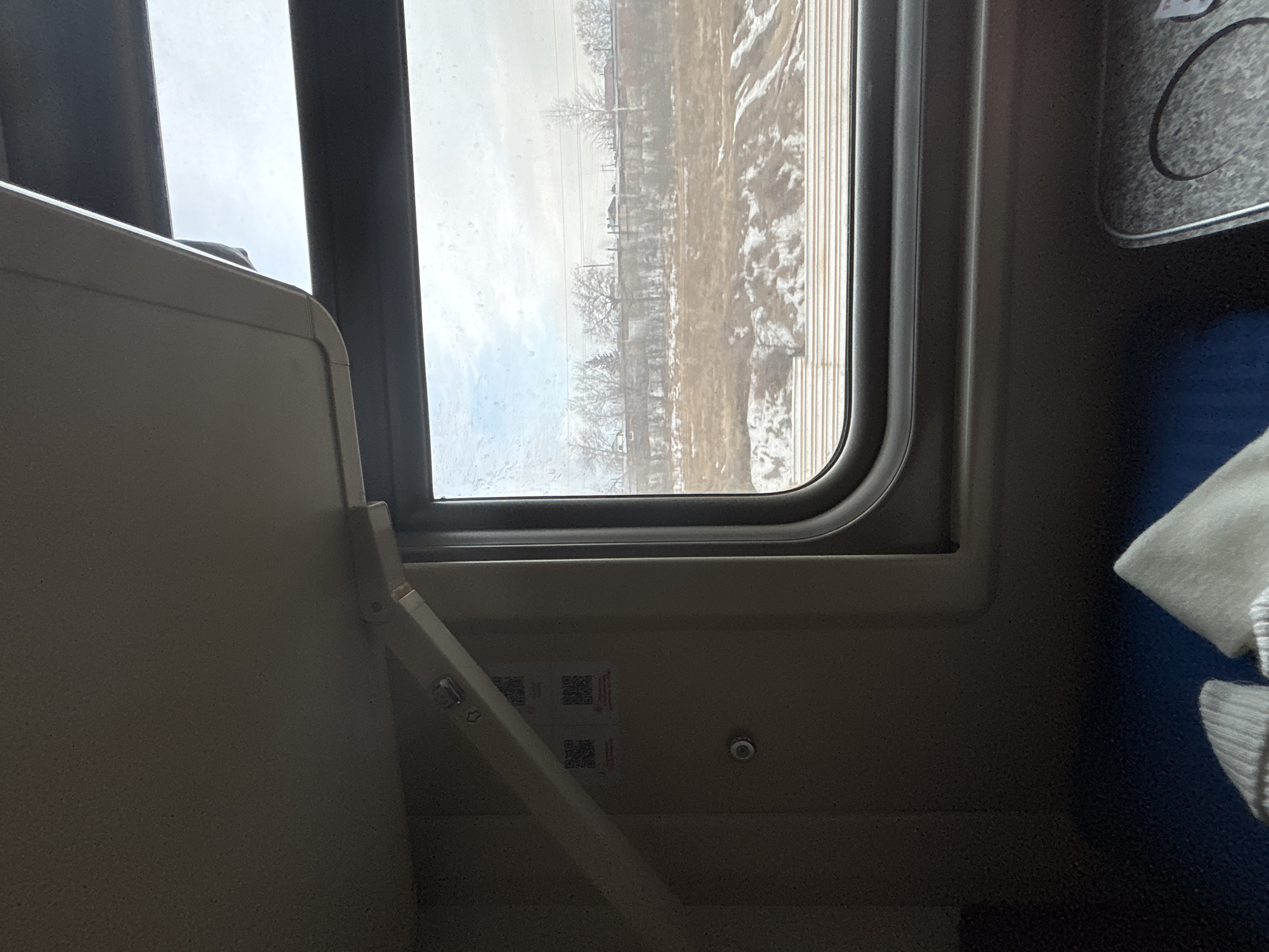 The image depicts the interior of a train car, observing an external winter landscape near Usolye-Sibirskoye, Russia, during daytime.
Inside the train, a light-colored, hinged overhead bunk or storage shelf is partially lowered, supported by a beige articulating arm. Below it, on a beige wall panel, two square QR codes with Russian text are affixed, positioned above a small, round silver button. A dark grey, rounded window frame encloses the view outside, its glass displaying visible smudges. In the lower portion of the image, a dark blue fabric surface, likely a seat or lower bunk, holds light-colored, knitted garments. To the bottom right, a silver-textured surface features a circular indentation, indicative of a cup holder or integrated table.
Outside the window, a winter scene unfolds under an overcast sky. Multiple parallel train tracks are visible in the immediate foreground, with patches of snow covering the ground between them. Beyond the tracks, a strip of dry, brown vegetation is interspersed with irregular snowdrifts. A long, low concrete wall spans the mid-ground. Above and behind this wall, leafless deciduous trees and some evergreen foliage are visible, with distant buildings and overhead utility lines appearing on the horizon. No people are visible within the frame.