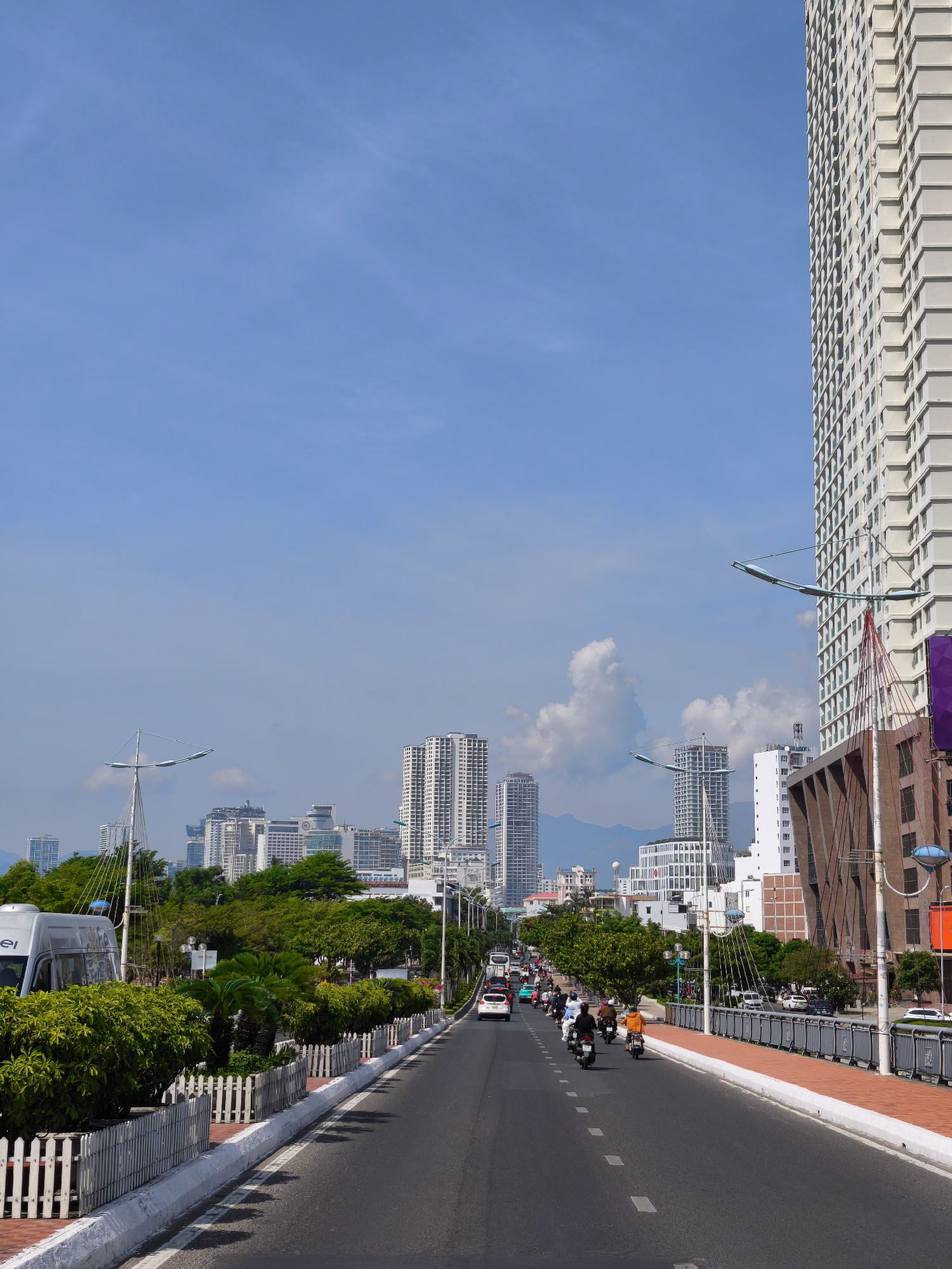 This is a vertical, eye-level shot of a busy road in a city. The sky is a clear, bright blue with a few wispy clouds. On the right side of the image, a very tall, modern white building dominates the skyline. Its facade is covered in a grid of windows and balconies. A bridge structure with red cables is visible in front of a building with a reddish-brown brick exterior. On the left side of the road, there are lush green trees and smaller buildings.
The road itself is paved and has white dashed lines marking the lanes. Numerous motorbikes and a few cars are traveling along the road, suggesting it's a bustling time of day, likely daytime given the bright sunlight and clear sky. The people on the motorbikes are dressed casually, wearing helmets. Some riders are wearing colorful clothing, like an orange shirt on one individual. The overall scene appears to be a typical urban environment with a mix of modern architecture and greenery. The presence of a bridge suggests the road might be elevated or crossing over water. There is no discernible text in the image.