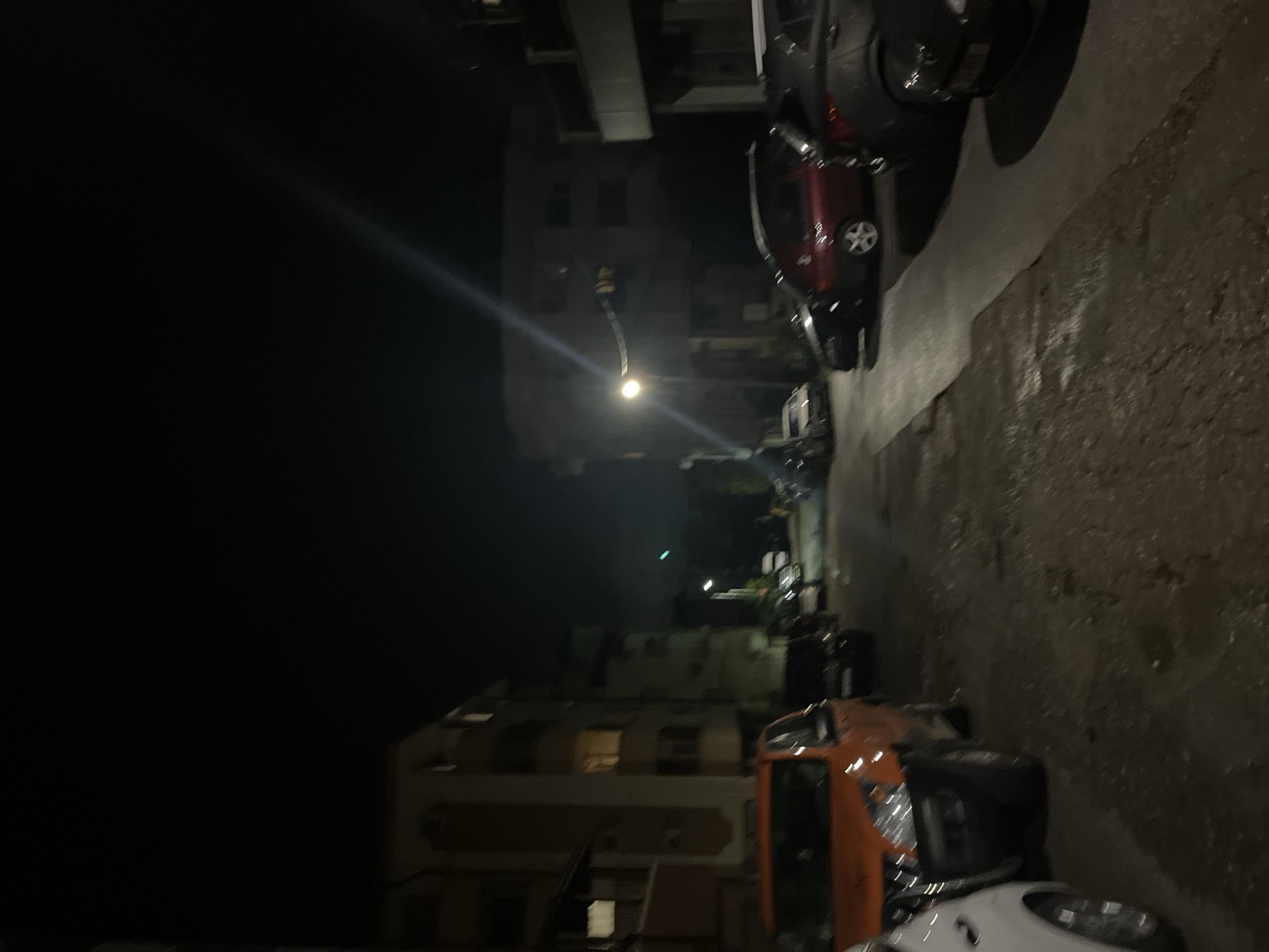 This is a nighttime street scene in Blagoevgrad, Bulgaria. The view is along a residential street with buildings on the left side and cars parked along the right side of the street. A street light illuminates the middle of the frame. The sky is dark and clear. The street is wet, suggesting recent rain. Parked cars vary in color from orange and white to red and dark gray.