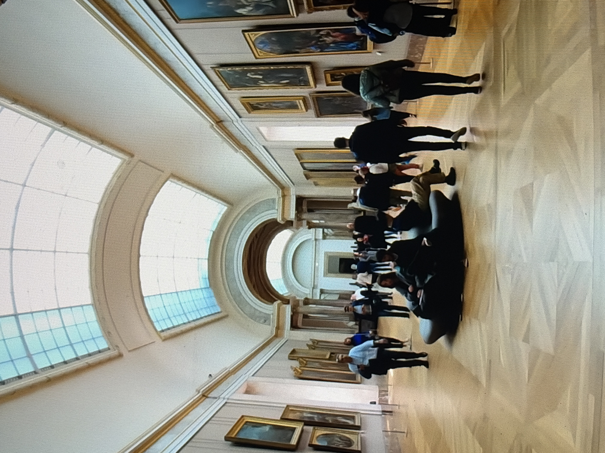 The image shows a long, wide museum gallery with a high, arched ceiling featuring a skylight. The walls are lined with numerous framed paintings, some large and ornate, others smaller. A polished wooden floor reflects the ambient light. A diverse group of people, dressed in casual attire, are walking and standing throughout the gallery, viewing the artwork. Some individuals are seated on a dark, curved bench in the foreground. The scene conveys a sense of public exhibition and visitor engagement within an art institution.