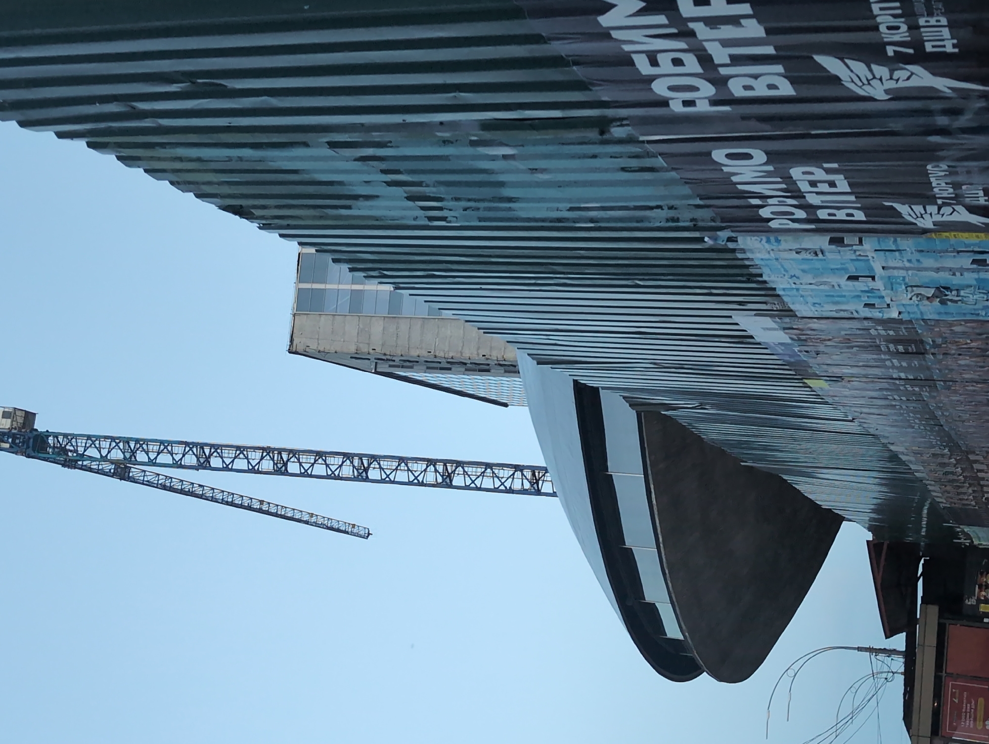 This is a ground-level, slightly upward-angled photograph of a construction site in Kyiv, Ukraine, on a day with a pale blue, overcast sky. A large construction crane with a blue boom dominates the upper left quadrant of the image. To the right of the crane, a modern, multi-story building with a glass facade is partially visible. In the foreground, a curved, dark-colored building with large windows is under construction, with a concrete-like texture. This structure is partially obscured by a tall, dark green, corrugated metal fence that runs diagonally across the right side of the frame. The fence is covered with numerous posters, some of which have Ukrainian text and graphics, including what appear to be stylized wings and the words "РОБИМО ВІТЕР" (meaning "We Make Wind" or "We Create Wind"). The lower left corner shows a street scene with a utility pole, wires, and what appears to be a small, red-roofed structure. The overall impression is one of ongoing urban development and construction.