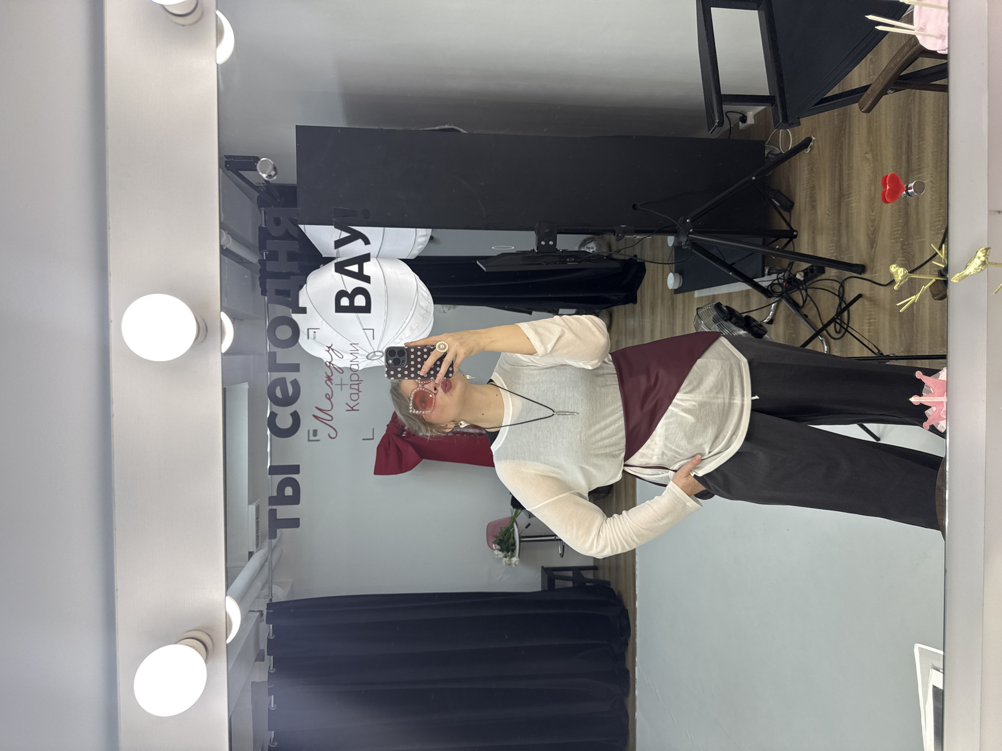 The image shows a woman in a room with makeup lighting, presumably in Izhevsk, Russia. The environment suggests a photo studio or dressing room with dark curtains, light wooden flooring, studio lighting equipment, and decor items such as decorative birds and flower arrangements. In the mirror's reflection, the woman holds a smartphone and appears to be taking a photo of herself. There is text and graphics on the mirror and on a large balloon, written in Russian. The woman's attire includes light-colored long-sleeved top, dark trousers and sunglasses. Her pose and the room setup suggest she is capturing an image of herself in a photo shoot or similar event.