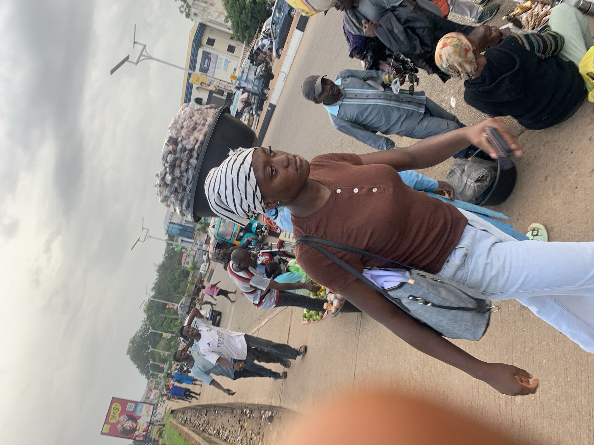 In the city of Barakin Lerawi, Nigeria, a woman wearing a brown t-shirt and a headscarf walks on a paved sidewalk. She carries a gray bag over her left shoulder. Behind her, a large black basin overflows with goods, likely produce. Several other individuals are visible on the sidewalk and in the street. To the left, two men in athletic wear walk along the sidewalk. Further back, a man in a white t-shirt with "Emirates" printed on it walks with his hand on his head. A person in a pink shirt walks in the background. On the street, a man in a gray and blue striped shirt appears to be interacting with a vendor who is selling fruit. A motorcycle with a sidecar is parked on the right, and other vehicles are present on the road. Buildings and streetlights are visible in the background, under a cloudy sky.