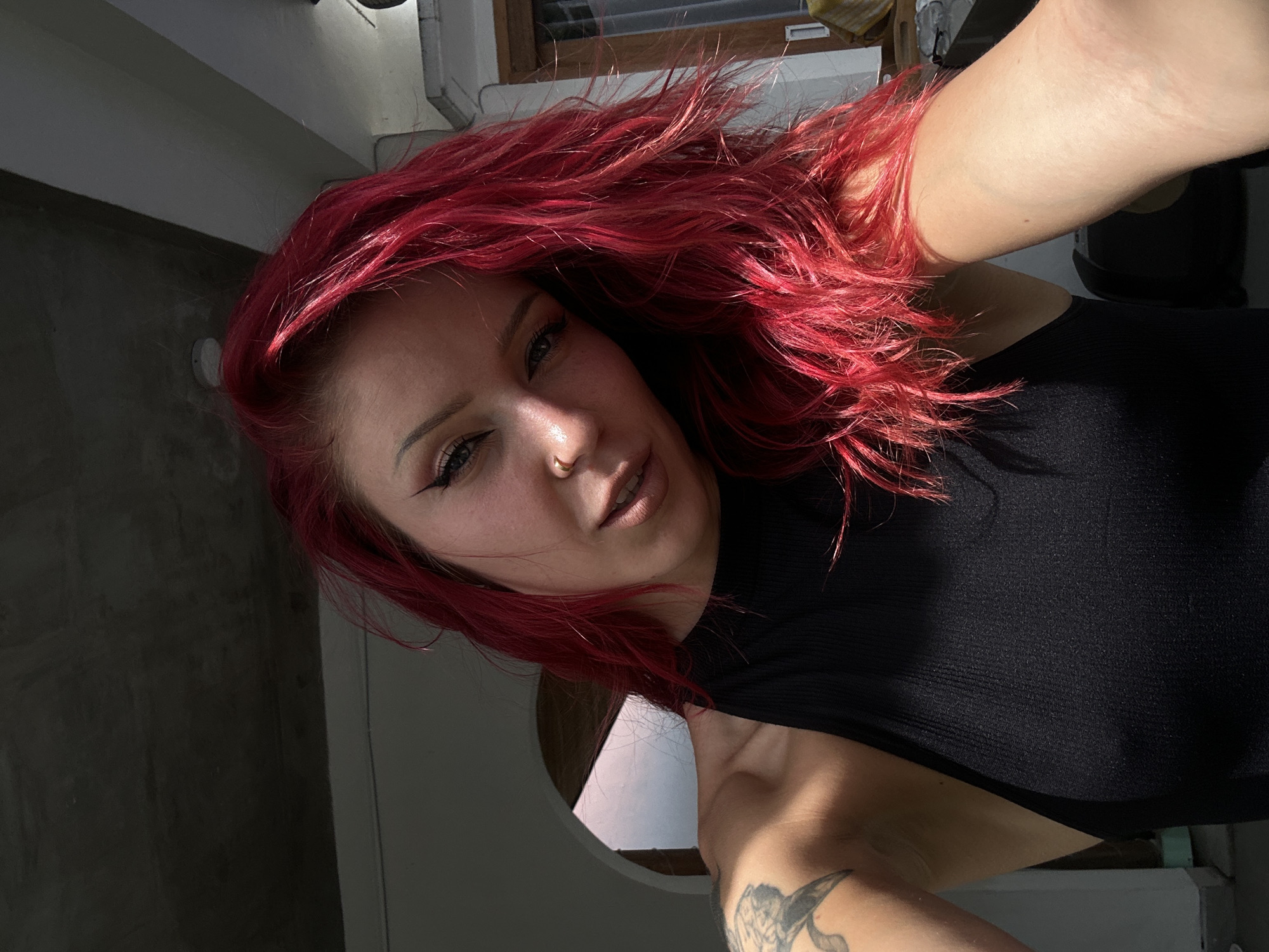 A fair-skinned woman with dyed red hair, wearing a black top, takes a selfie at an indoor location, possibly in North Kuta, Indonesia, based on visual cues and the query. The woman has a nose ring and visible eyeliner. A tattoo is present on her arm. A window is visible in the background.