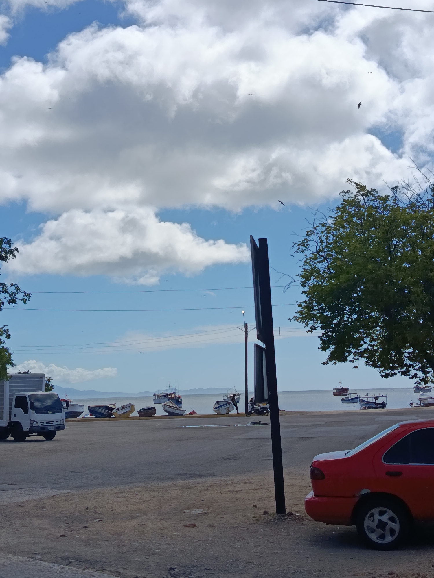 The image shows a view of the Juan Griego bay in Venezuela. In the foreground, a red car is parked on a gravel lot, with a large, dark pole to its left. To the left of the pole, a white truck is parked. In the middle ground, several small, colorful fishing boats are pulled up onto the shore, some tilted on their sides. The water of the bay is calm, and in the background, a hazy landmass with mountains is visible under a bright blue sky dotted with fluffy white clouds. Several birds are visible flying in the sky.