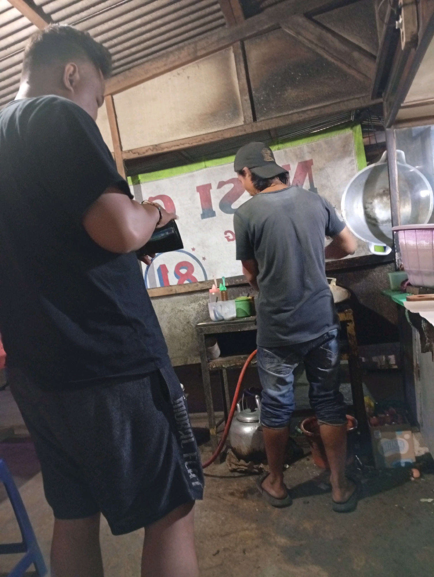 In Caringin, Indonesia, an outdoor scene depicts two men. The man on the left, facing away from the camera and slightly to the right, wears a black t-shirt and dark shorts. He holds a black object, possibly a wallet or phone, in his left hand. The man on the right, also facing away, wears a dark grey t-shirt, blue jeans, and a black baseball cap. He is positioned near a counter with various items, including a large metal pot or basin, a red hose, and several containers. The environment appears to be a food stall or eatery, with a corrugated metal roof overhead and a banner with lettering visible on the wall behind the men. The ground is concrete.