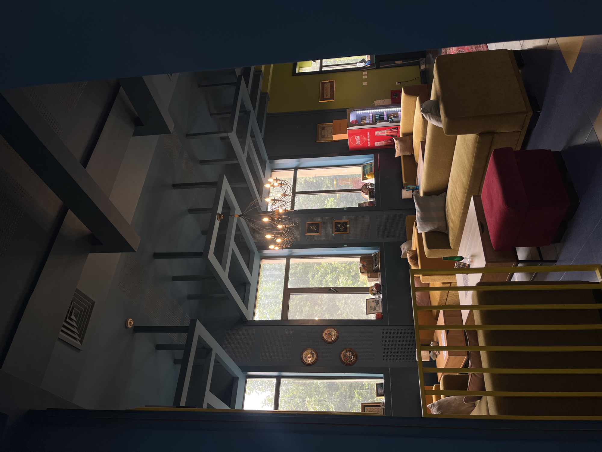 The image displays the interior of an unoccupied cafe or lounge area, bathed in natural daylight. The view is from a slightly elevated position, looking across a spacious room.
The dominant features on the right side of the frame are several mustard yellow sofas and armchairs, arranged with wooden coffee tables in front of them. Cushions in various light and dark shades are placed on the seating. A red upholstered ottoman or cube seat adds a vibrant accent. These seating arrangements are partially obscured by yellow metal railings, suggesting a mezzanine or balcony from which the photo was taken. The floor in this area has a geometric pattern of dark blue and yellow or light blue tiles.
The walls are primarily a dark blue-gray, with the left wall and ceiling featuring a distinctive geometric design of protruding dark gray beams or frames. A multi-bulb chandelier hangs from the ceiling, providing additional light. Large windows along the back and left walls allow abundant sunshine to enter, showcasing lush green trees and foliage outside, indicating a bright daytime setting.
In the background, a section of wall is painted olive green, against which a red "Coca-Cola" refrigerator is visible, stocked with beverages and displaying the slogan "TASTE THE FEELING". Various framed pictures, including ornate plates, adorn the walls. A small, vintage-style typewriter can be seen on a shelf near a window. An air vent is embedded in the ceiling.
The absence of people contributes to a serene and inviting atmosphere. The mix of warm yellows and cool blues, alongside modern architectural elements, creates a unique and stylish interior.