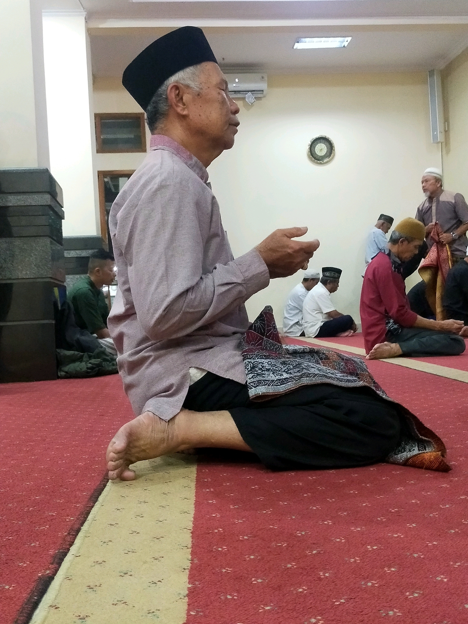 The image depicts an indoor scene, likely a prayer hall or mosque, with several men engaged in prayer or religious activities. The primary subject is an elderly man in the foreground, wearing a black peci (a cap) and a light-colored, long-sleeved tunic over black pants. He is kneeling on a red carpet with a yellow stripe, his right foot bent at the ankle and his left leg tucked underneath. His hands are raised in prayer.
In the background, other men are also visible, similarly dressed in traditional Islamic attire. One man in a maroon outfit and a yellow peci is kneeling to the right of the main subject. Further back, several other men are seated or standing.
The environment appears to be well-lit, with fluorescent lights visible on the ceiling. A clock is mounted on the wall, and what appears to be an air conditioning unit is also present. The flooring is a red carpet with a subtle pattern, and there are some dark gray stone structures on the left side of the image, possibly part of the building's architecture. The overall atmosphere suggests a religious gathering. The image does not provide specific details about the city of Bandung City, Indonesia, or any precise location within it.