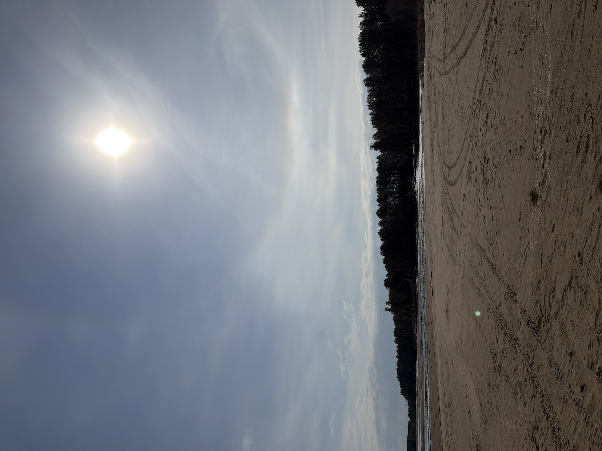 The image shows a landscape in Chaikovsky, Russia, featuring a sandy beach with tire tracks, a dark line of trees in the distance, a pale sky, and the sun. The beach is brown, and wet in some parts. There is no one visible in the image.