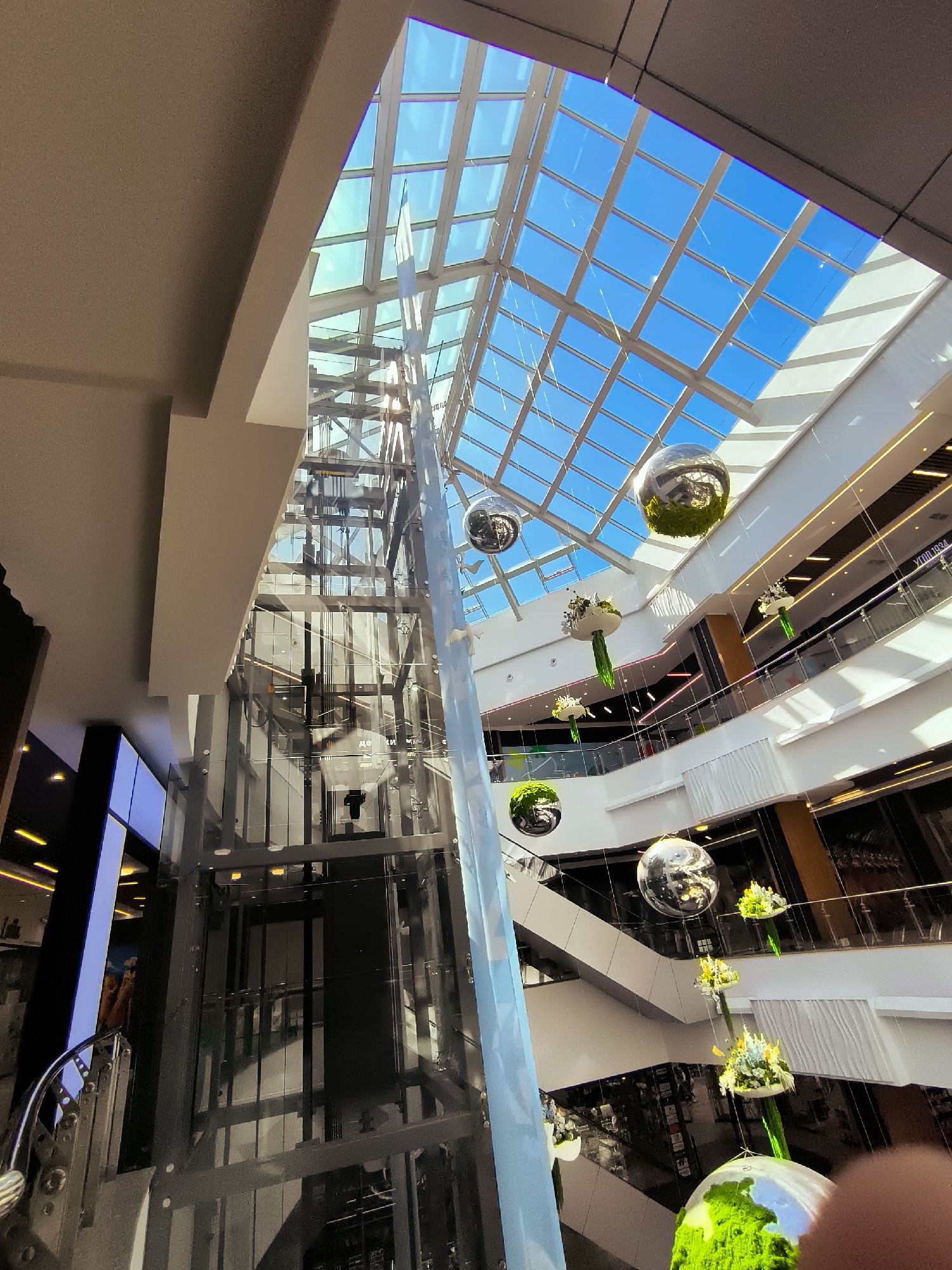 The image depicts the interior of a multi-level shopping mall with a distinctive glass ceiling that allows natural light to illuminate the space. A prominent glass elevator shaft is visible on the left side of the frame, with the elevator car itself at a lower level. Several decorative spherical orbs, some appearing to be covered in moss or greenery, are suspended from the ceiling by thin wires, along with smaller floral arrangements. The mall features multiple levels with balconies and railings, and shops or businesses are visible on these levels. The overall impression is one of a modern, well-lit commercial space with artistic decorations. The provided context that this location is in Qarağandı city administration, Kazakhstan, places the scene within an urban environment.