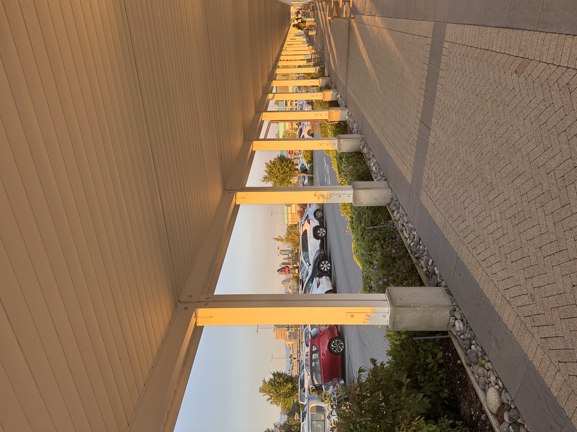 The image depicts a covered walkway with a white, corrugated ceiling and square concrete pillars spaced evenly apart. The walkway runs alongside a parking lot filled with various vehicles, including cars and SUVs. Beyond the parking lot, a city skyline with buildings and mountains in the distance is visible under a clear sky bathed in the warm light of sunset. The ground in the foreground of the walkway is paved with beige interlocking bricks arranged in a herringbone pattern, transitioning to a darker grey band. Adjacent to the parking lot is a landscaped area with green bushes, flowers, and decorative rocks. In the distance, on the walkway, two individuals are walking, one pulling luggage. The scene appears to be in the city of Richmond, Canada, likely a commercial area or transit hub given the parking lot and pedestrian walkway.