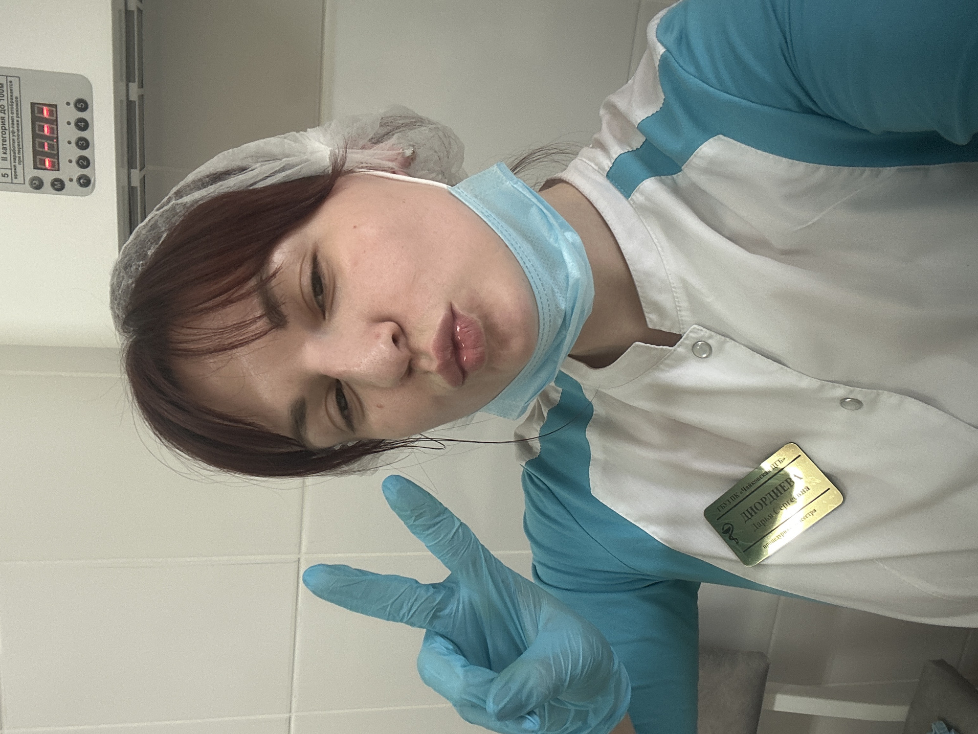 A young woman is posing for a selfie in what appears to be a medical setting. She is wearing a white and blue medical uniform, a blue surgical mask around her neck, and a disposable hairnet. She is wearing a blue medical glove on her right hand and holding up two fingers in a peace sign. Her face is turned to the side, and she is pursing her lips. A gold-colored name tag pinned to her uniform reads "Диордиев, Дарья Сергеевна," along with the text "ГБУЗ ПК 'Чайковская ЦГБ'" which refers to a medical institution. The text below her name reads "процедури сестра." A piece of equipment is visible in the background, with the text reading "ІІ категория до 100М" and "время наработки уф-ламп отображается при переключения режимов." The equipment's display shows the numbers "8.8.8.8." The setting appears to be a tiled room. The mentioned "Чайковская ЦГБ" is in Чайковский, Russia, which implies the location of the image is also somewhere near the city of Чайковский, Russia, possibly even a district in the city like Фоки.