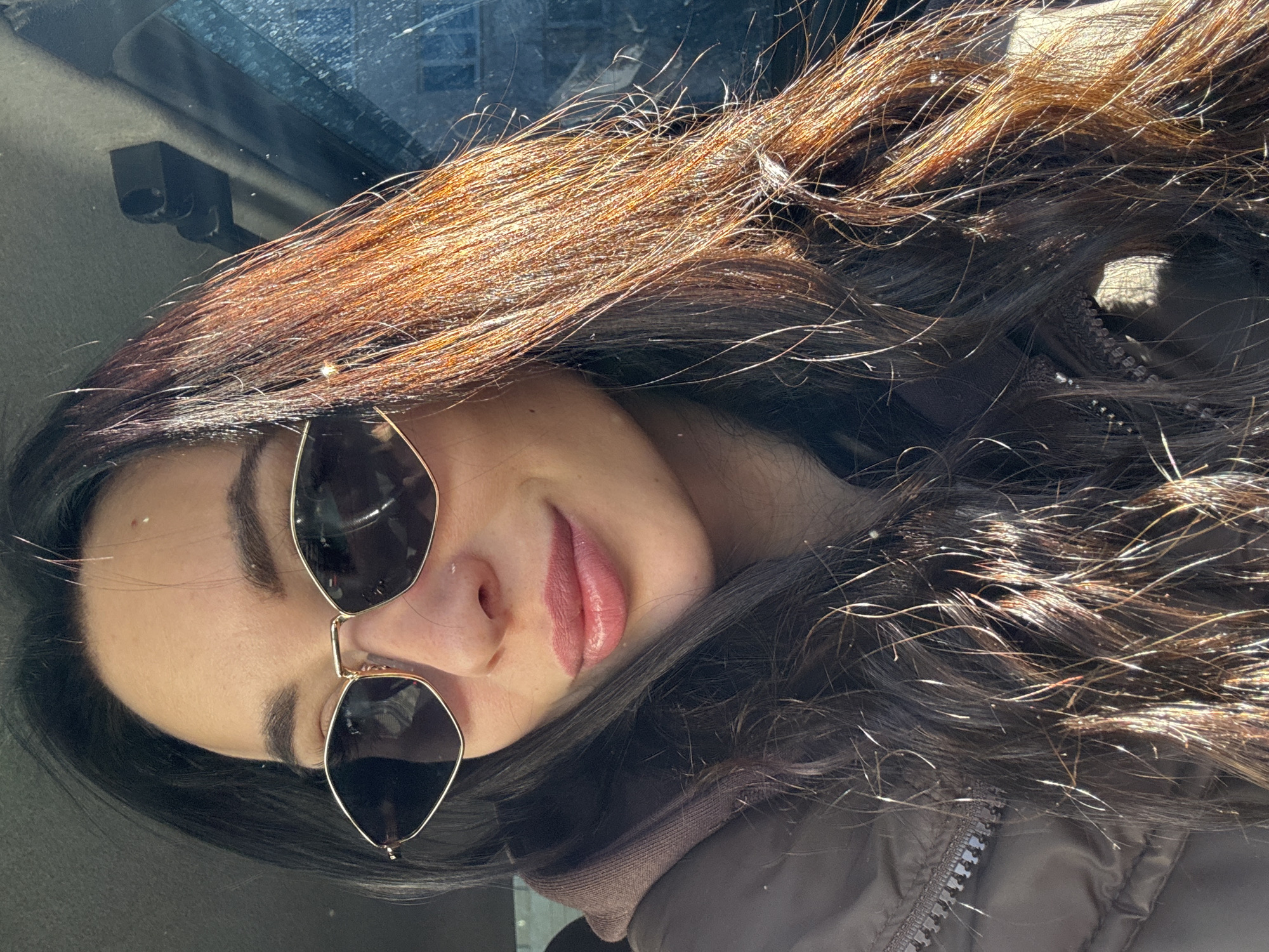 A young woman is posing for a selfie in what appears to be a car. She has long, dark hair with lighter brown highlights, and is wearing sunglasses with gold frames. Her lips are slightly parted. She is wearing a brown, possibly quilted, jacket. The interior of the car is visible in the background, including a rearview mirror. There is no clear evidence in the picture that suggests she is in the city of Chaikovsky, Russia.