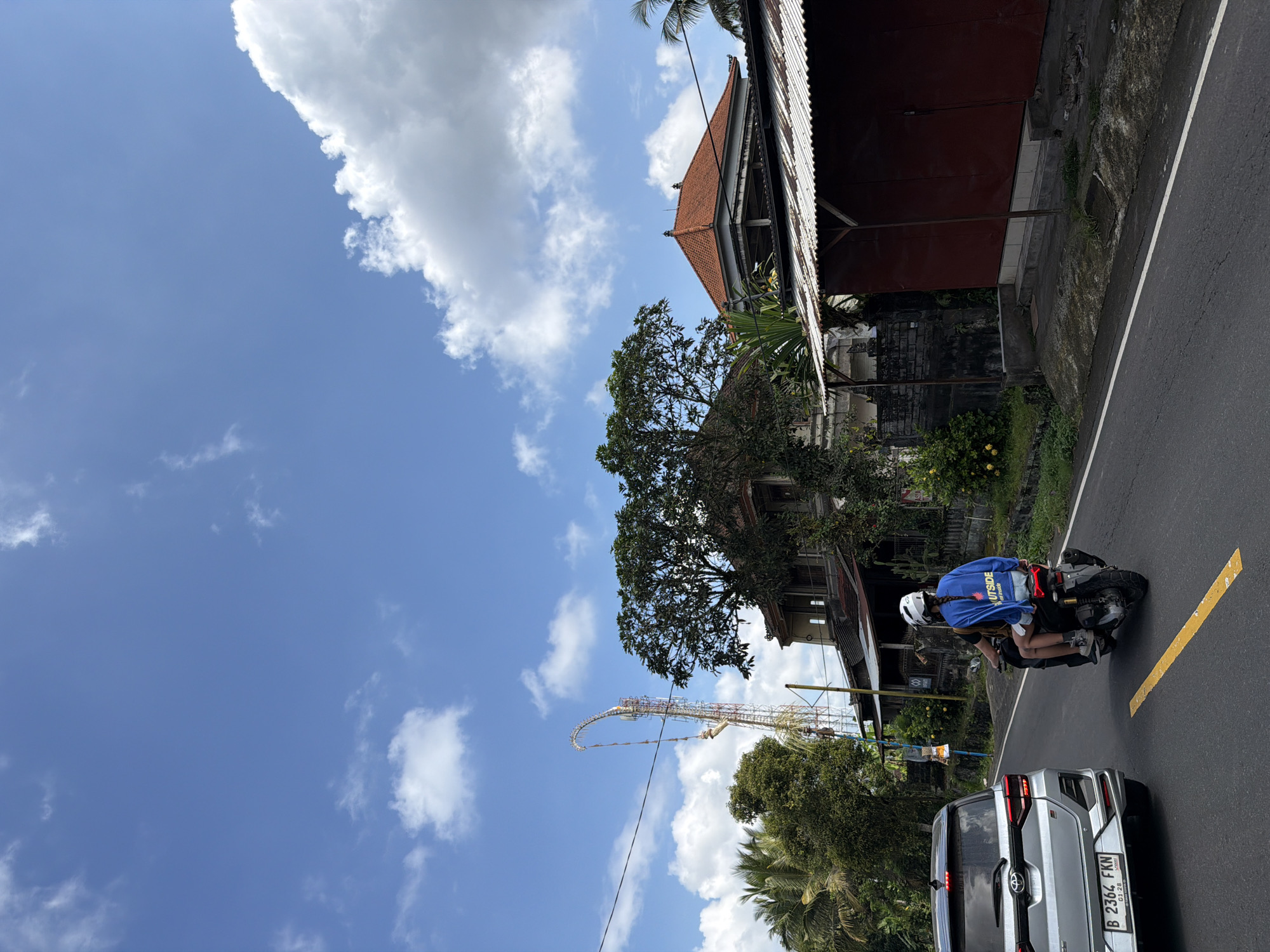 The image depicts a street scene in Tampaksiring, Indonesia, under a bright blue sky with scattered white clouds. A silver SUV is visible on the left side of the asphalt road, displaying a license plate that reads "B 2364 FKN". Further down the road, two people are riding a scooter. The person in the back is wearing a blue sweatshirt with the word "OUTSIDE" printed on it. A tall, curved structure, possibly a telecommunications tower, is prominent in the background, with buildings and lush green trees surrounding it. The architecture of the buildings suggests a tropical location, with red tiled roofs and open-air structures. The overall atmosphere is that of a sunny day in a bustling town or village.