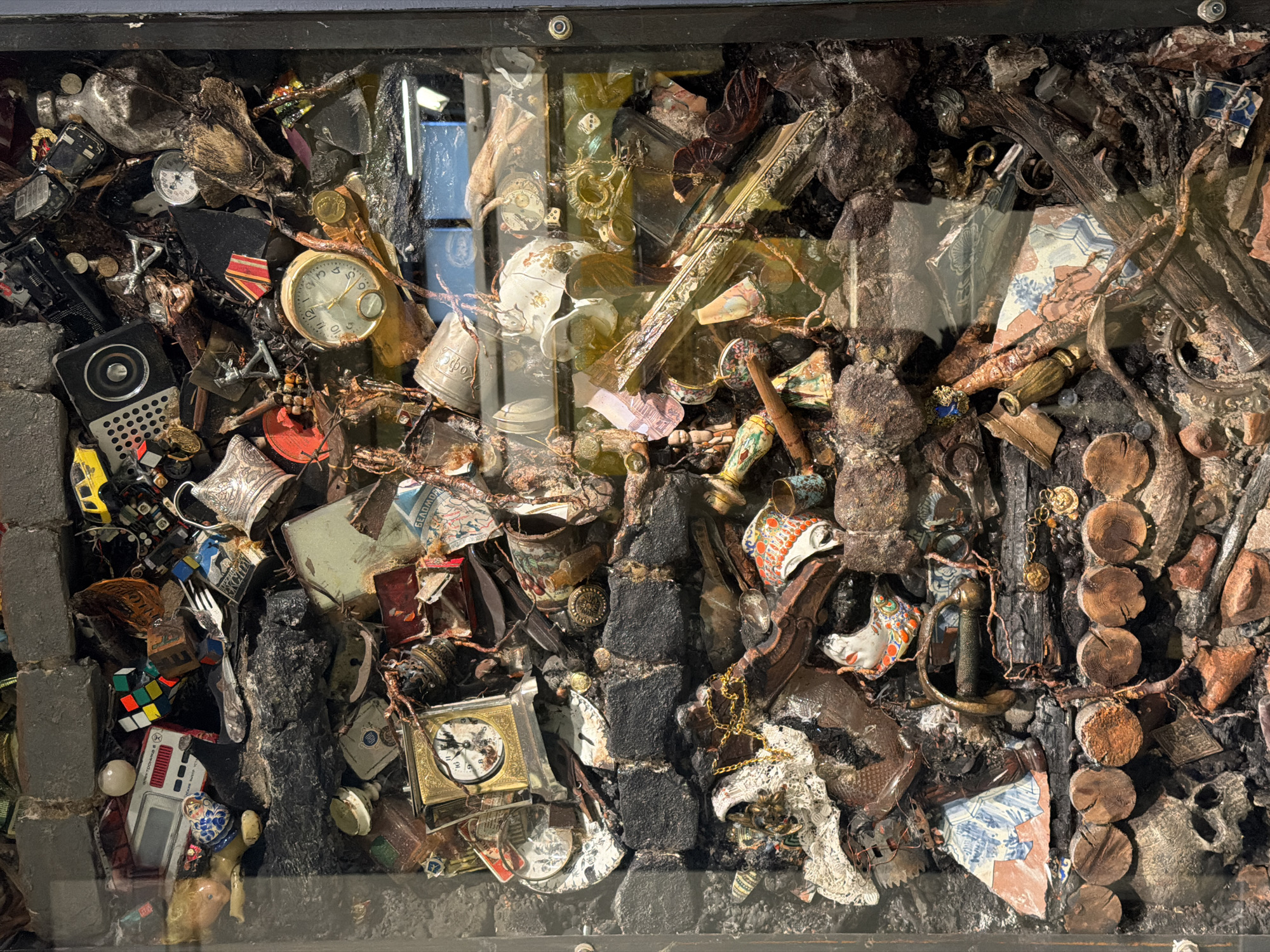 This is a close-up photograph of an art installation or display in what appears to be a glass-covered container or vitrine. The artwork is composed of a densely packed collection of various objects and materials, including broken porcelain, clock parts (faces and hands), metallic objects, fragments of decorative frames, wood pieces, and other assorted items. The arrangement has a chaotic, almost archaeological, feel, suggesting a collection of salvaged or found objects. The overall color palette is muted, with earth tones, grays, and metallic hues dominating, although there are pops of color from the porcelain fragments and other items. Due to a lack of visual clues, it is not possible to confirm that this piece is located in Saint Petersburg, Russia.