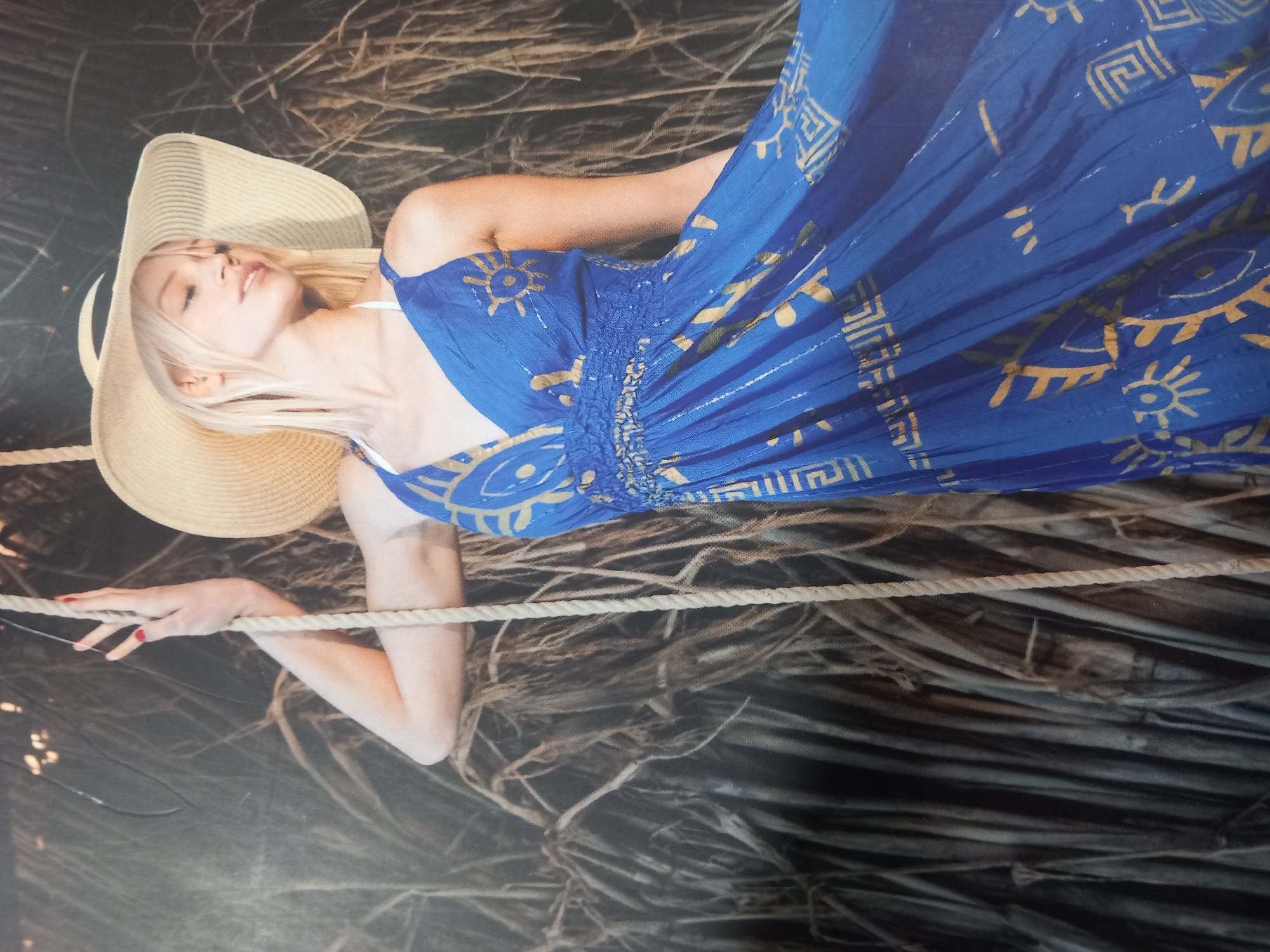 A young, blonde woman with fair skin, wearing a wide-brimmed straw hat and a flowing blue dress with a gold pattern, leans against a rope. The dress has a V-neck and a smocked waist. Her right arm is extended upwards, with her hand resting on the rope. The background appears to be a dark, textured wall or a dense arrangement of dried foliage. The lighting is soft, highlighting the woman and her attire. The location is unknown.