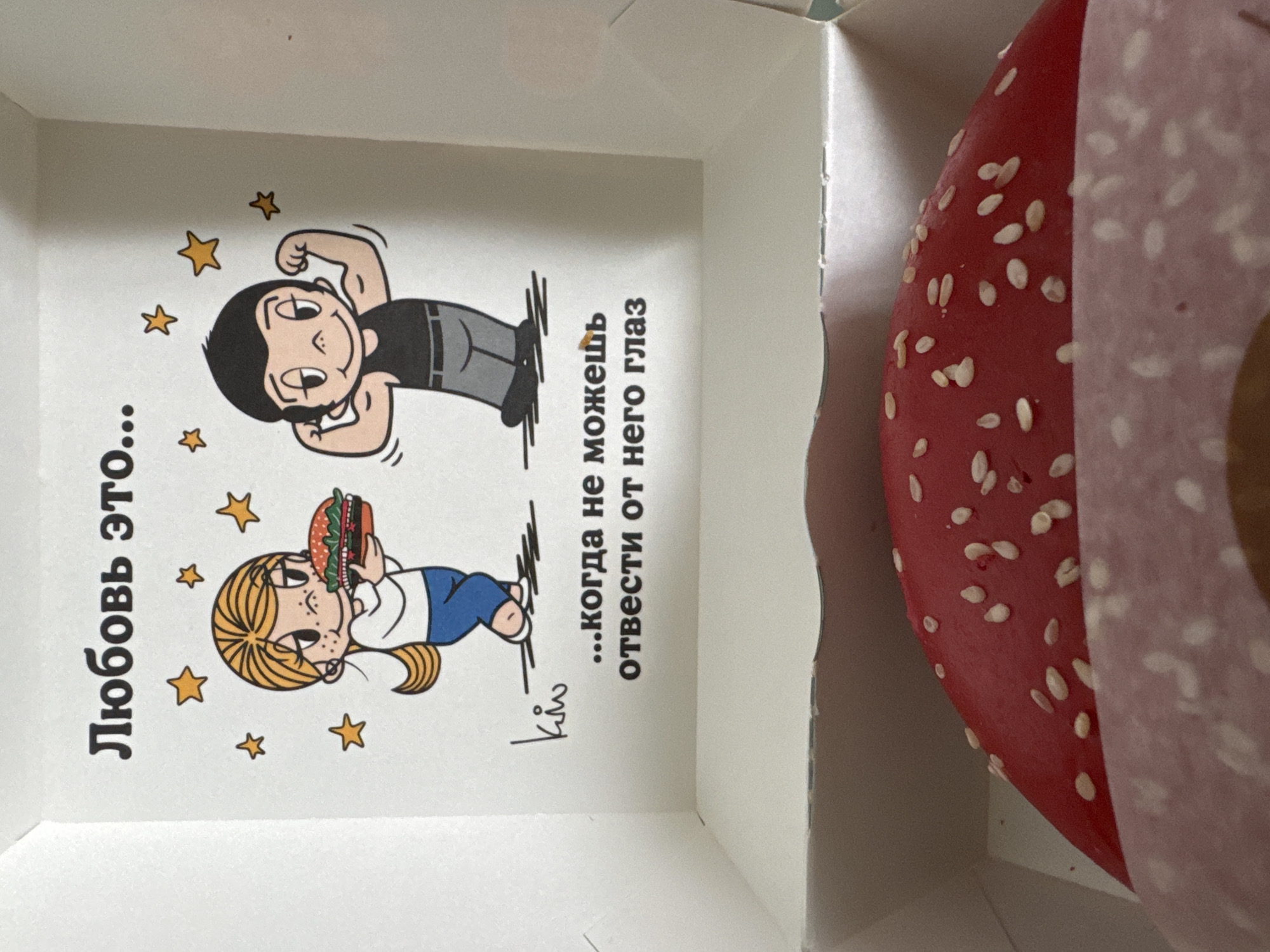 The image shows the interior of a white cardboard food box. On the left side of the box, there's a cartoon drawing of a woman holding a burger and a man flexing his muscles. The drawing is accompanied by Russian text that reads "Любовь это... ...когда не можешь отвести от него глаз," which translates to "Love is... ...when you can't take your eyes off of him/her." The drawing is also surrounded by cartoon stars. On the right side of the box is a red burger bun with white sesame seeds. It is partially covered with a white paper wrap.