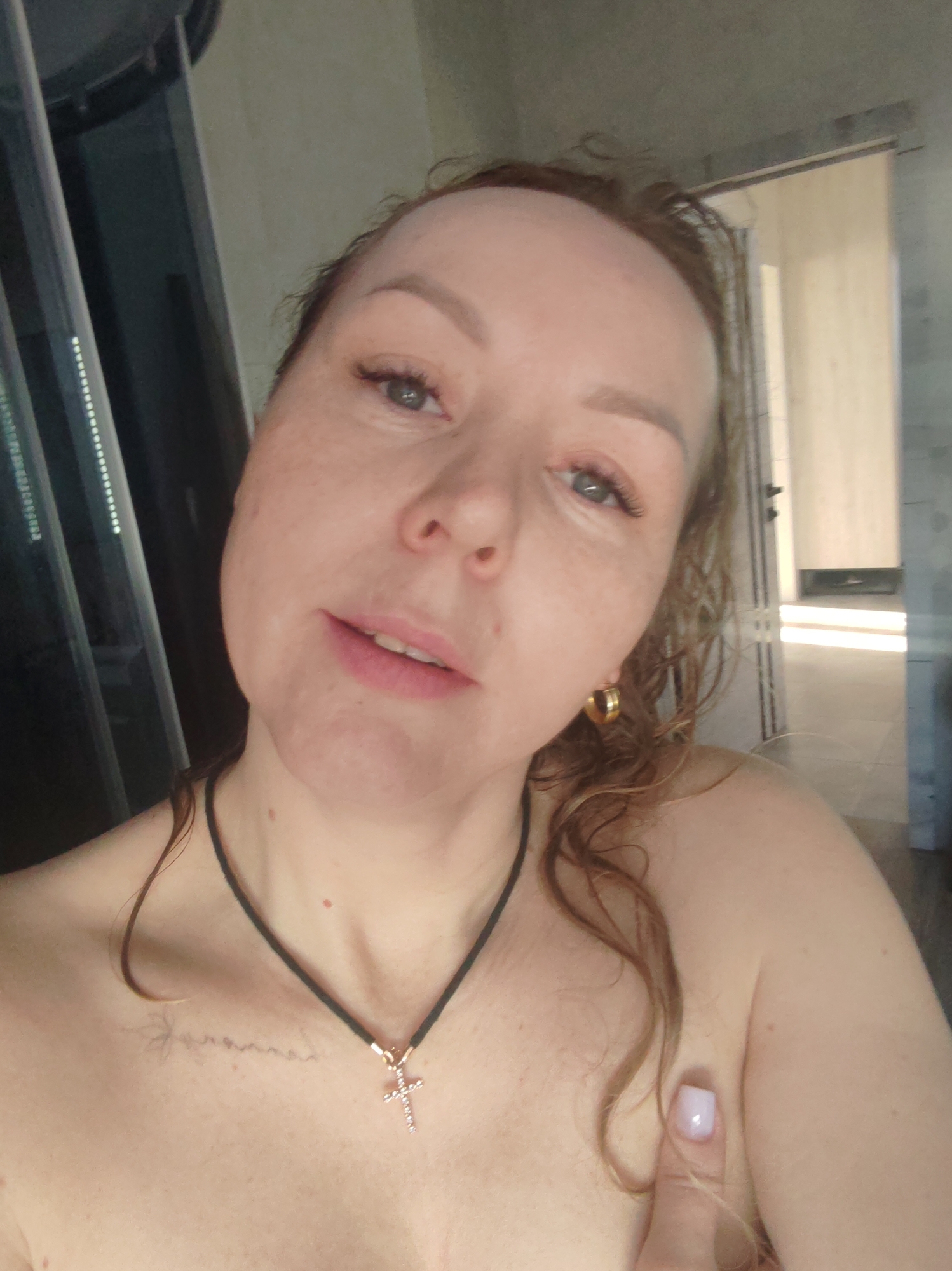 A selfie-style photograph depicts a woman with fair skin and light brown hair, likely in an indoor setting. She has freckles, light-colored eyes, and a gold hoop earring visible in her left ear. She is wearing a black choker necklace with a small cross pendant. A faint tattoo is noticeable on her chest, partially obscured by a strand of her hair. The background suggests a bright interior with doorways leading to other rooms, possibly a residential building. It is not possible to identify the location as Chaikovsky, Russia from the given image.