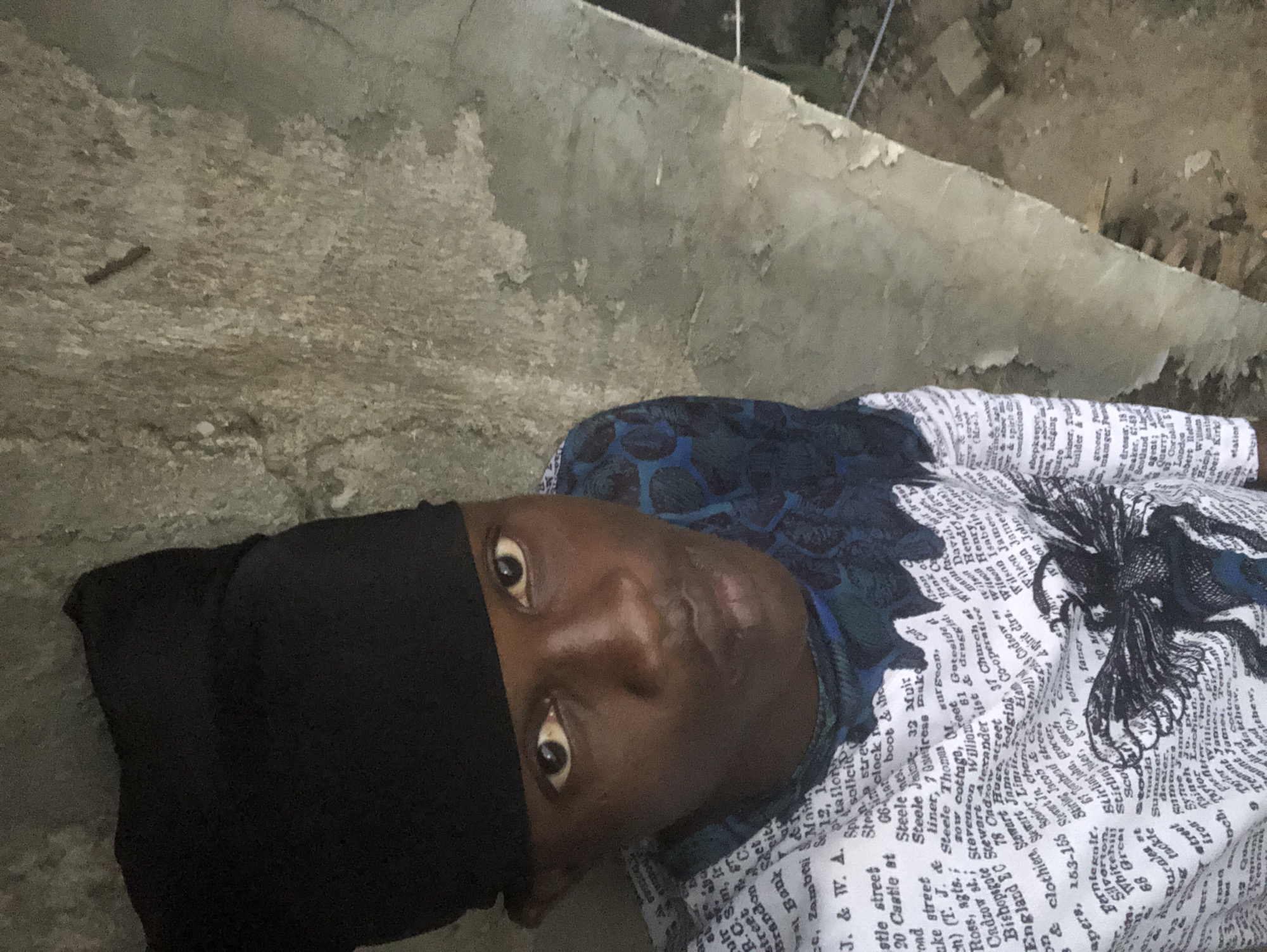 A young Black male wearing a black head covering and a white t-shirt with text and a dark graphic is standing against a textured, weathered concrete wall. The lighting appears to be natural, casting shadows and highlighting the texture of the wall. The man is looking towards the camera with his mouth slightly open. The background shows a rough, uneven concrete surface, suggesting an outdoor or unfinished urban environment, possibly in Ado, Nigeria, given the context of similar images from that region. The wall has an aged appearance with peeling paint or plaster.