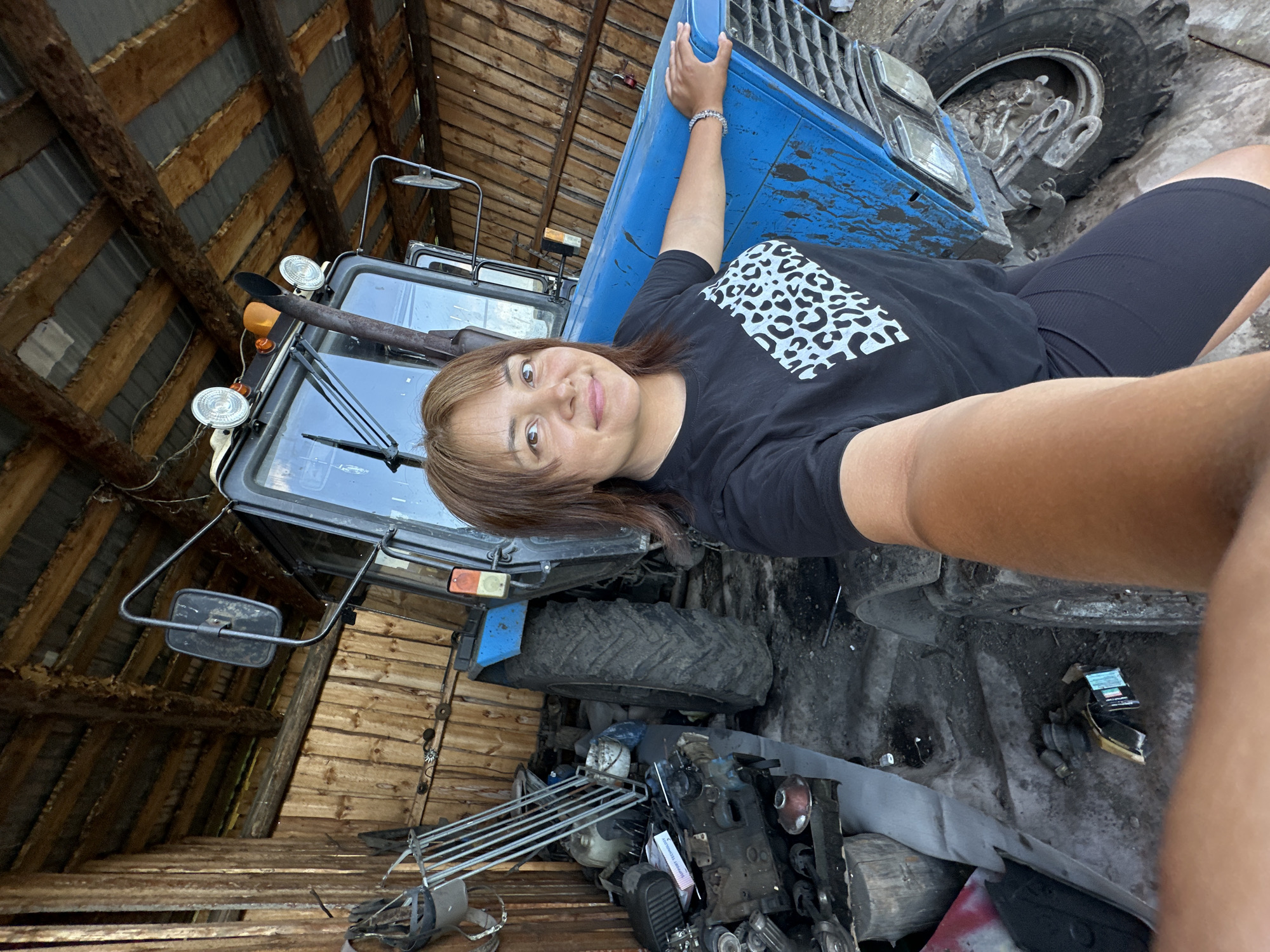 A woman is taking a selfie in a barn. She is wearing a black t-shirt with a leopard print design and black shorts. She is leaning on the hood of a blue tractor. The barn has a wooden roof and walls. There are various tools and parts of machinery scattered around the barn. The ground is dirt and concrete.