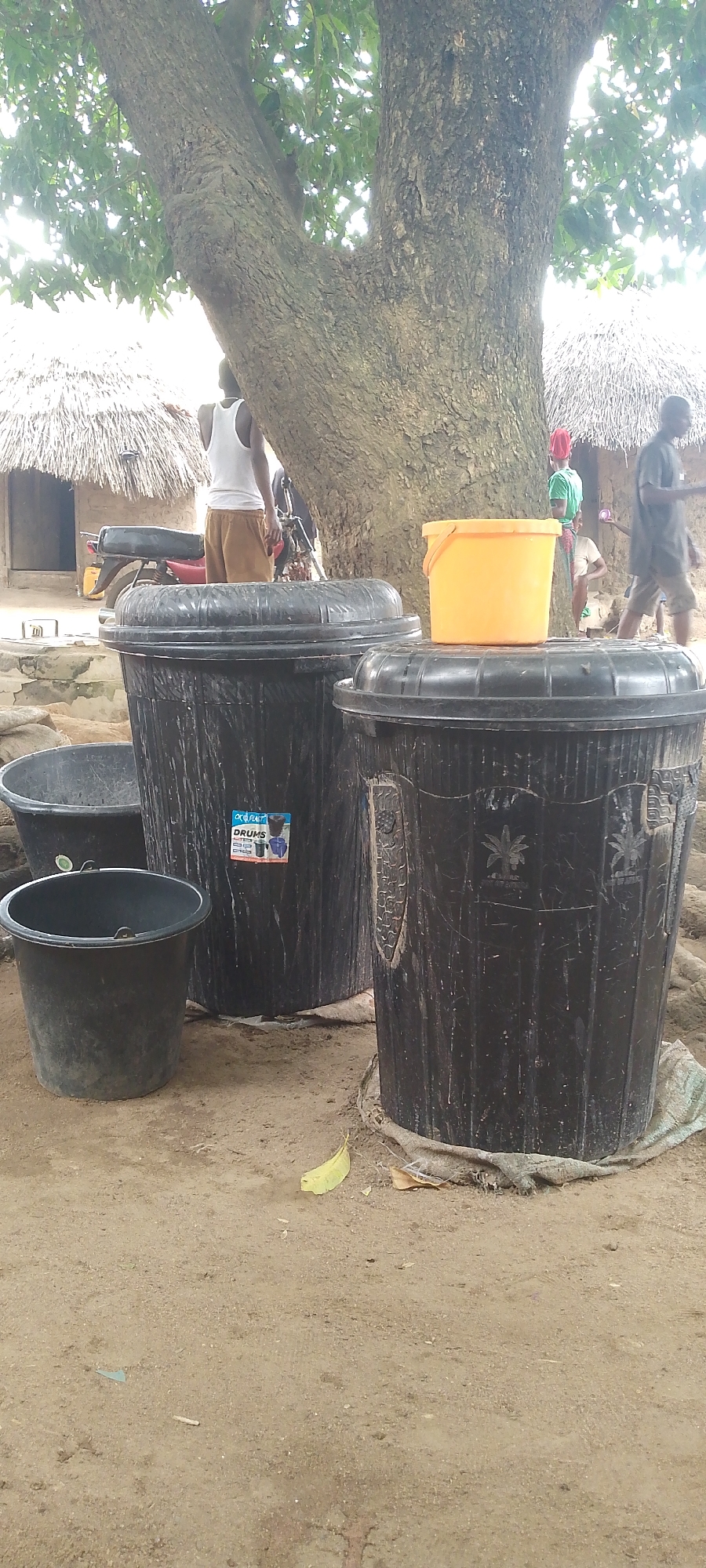 An outdoor scene shows several large black plastic drums and two smaller black plastic buckets in the foreground, arranged on sandy ground. One of the large drums has a yellow plastic bucket placed on top of it. A man wearing a white tank top and brown shorts stands behind the drums. In the background, a thatched-roof building and a motorcycle are visible. Several people are standing in the distance. The scene is dominated by a large tree with green leaves.
