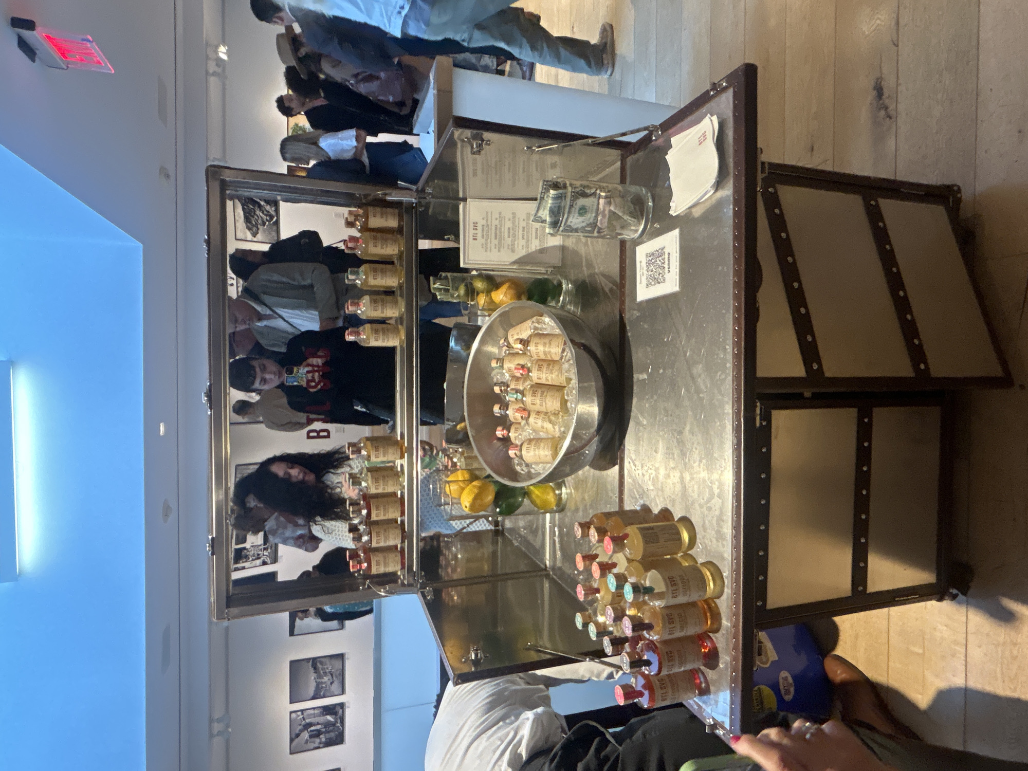 The image captures an indoor event featuring a portable bar or display stand, resembling an open, metal-clad trunk. The stand is well-stocked with numerous small bottles, likely containing beverages like spirits or cocktails. Many bottles are stacked neatly, while others are presented in a bucket filled with ice, alongside lemons and limes.
On the stand's surface, a glass jar serves as a tip container, holding cash bills. A white card, possibly a menu, and a QR code sticker with the "Venmo" logo are also visible. In the background, several people are gathered, some interacting with the stand, while others mingle further back. The setting appears to be an event space or gallery, with black and white photographs adorning the walls. A red "EXIT" sign is visible near the ceiling.
The overall scene suggests a social gathering, possibly a tasting event or beverage service. The lighting is bright and artificial, common in indoor venues. Visible text on the bottle labels includes "BTL SVC" and "DAS 118". A person's hand is visible in the bottom right, potentially engaging with the stand.