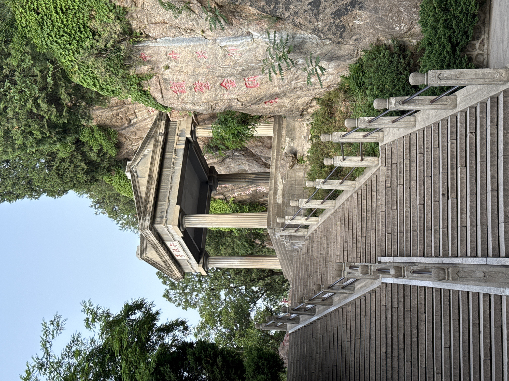 A stone staircase leads up to a small pavilion built against a rock face covered in red Chinese characters. The location is within the Lintong District of China. The scene includes greenery such as vines and trees surrounding the stone structures. The staircase has metal railings along the sides, and the pavilion has classical architectural elements with columns. The sky is visible in the background.