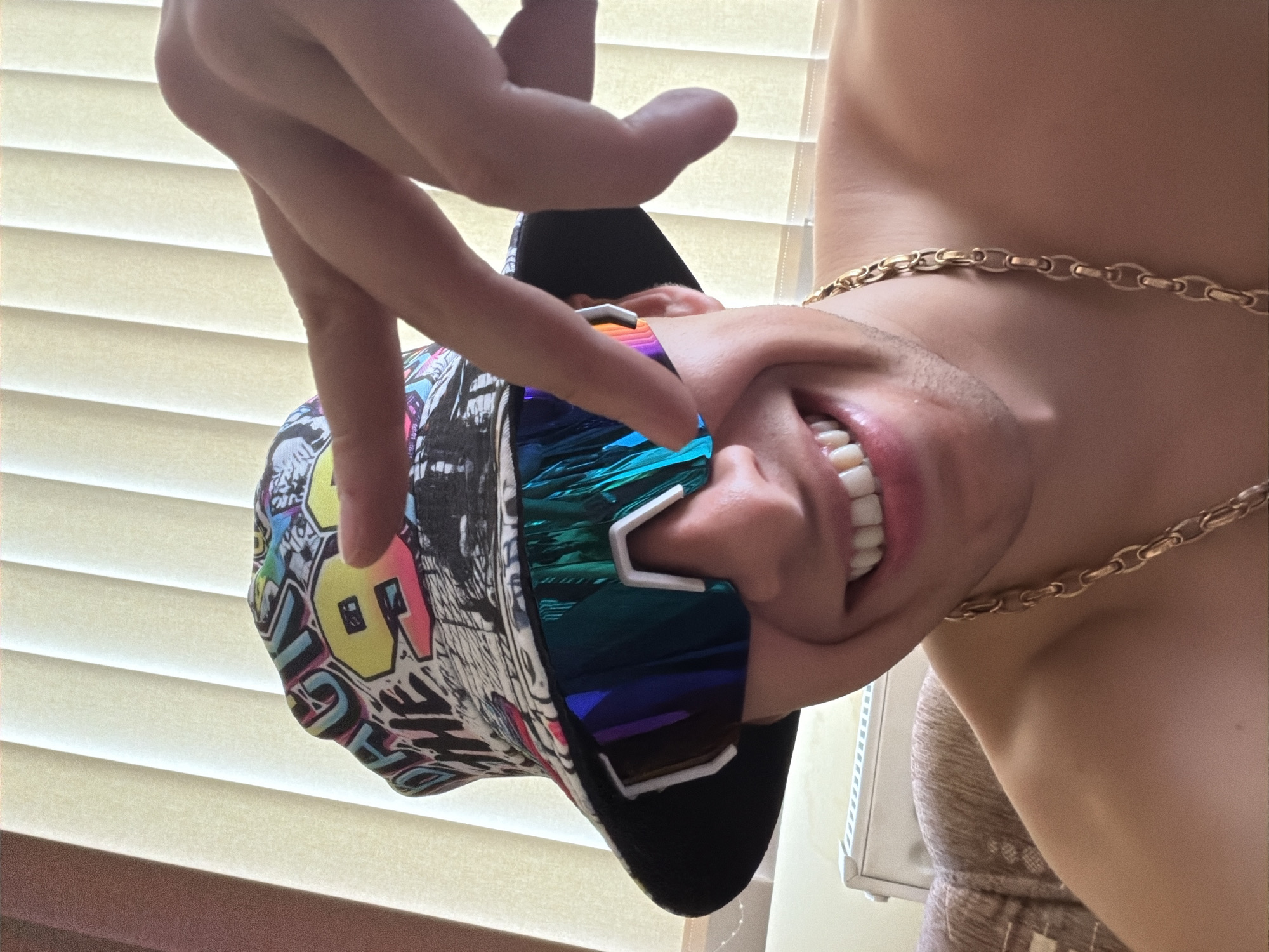 An upside-down, close-up shot captures a person's head and upper torso, illuminated by natural light filtering through horizontal window blinds in the background. The person is wearing a multi-colored cap adorned with graffiti-style patterns, including a prominent "LOVE" in yellow, pink, and light blue. Over their eyes are futuristic, mirrored sunglasses with white frames, reflecting vibrant blue, green, and purple hues. A gold-colored chain is draped around their neck and shoulder. The person is smiling broadly, revealing their teeth, with their mouth open in what appears to be a laugh or joyful expression. One hand is visible near their face, with fingers seemingly touching or adjusting the cap and sunglasses. The image is rotated, creating a dynamic and playful composition, suggesting the person might be hanging upside down or performing an inversion. The setting is indoors, likely by a window, during the daytime.