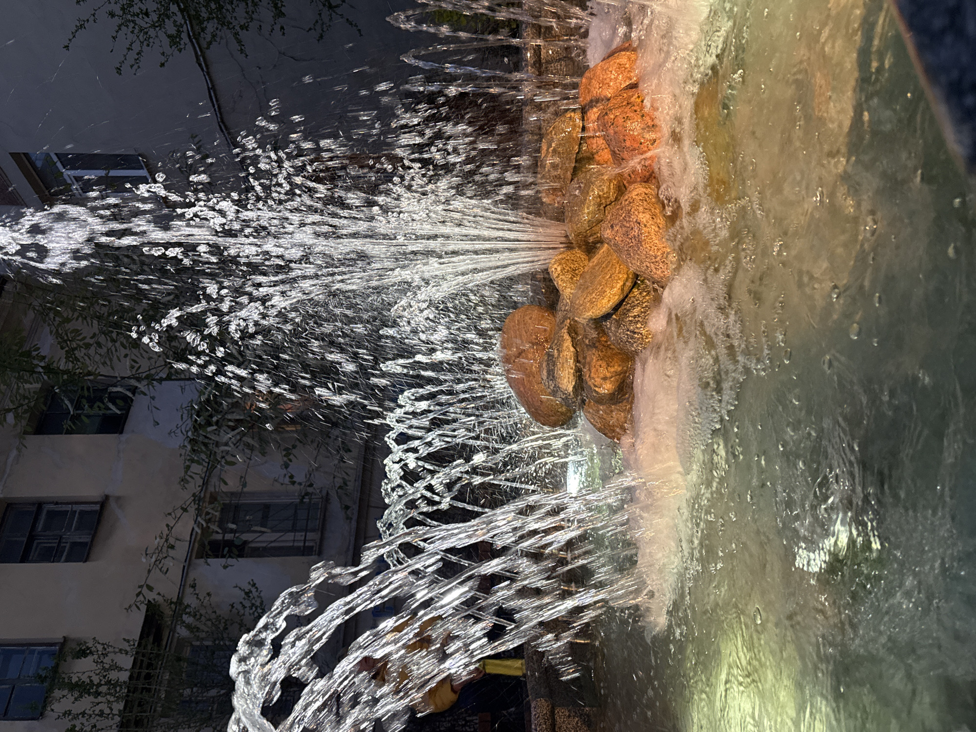 The image shows a fountain at night in Saint Petersburg, Russia. The fountain features a cascade of water jets spraying from a structure built with large, smooth stones. The water is illuminated, creating a sparkling effect. Behind the fountain, a light-colored building with dark windows is visible. Some greenery is also visible around the fountain. The overall scene is captured with a shallow depth of field, emphasizing the fountain while blurring the background.